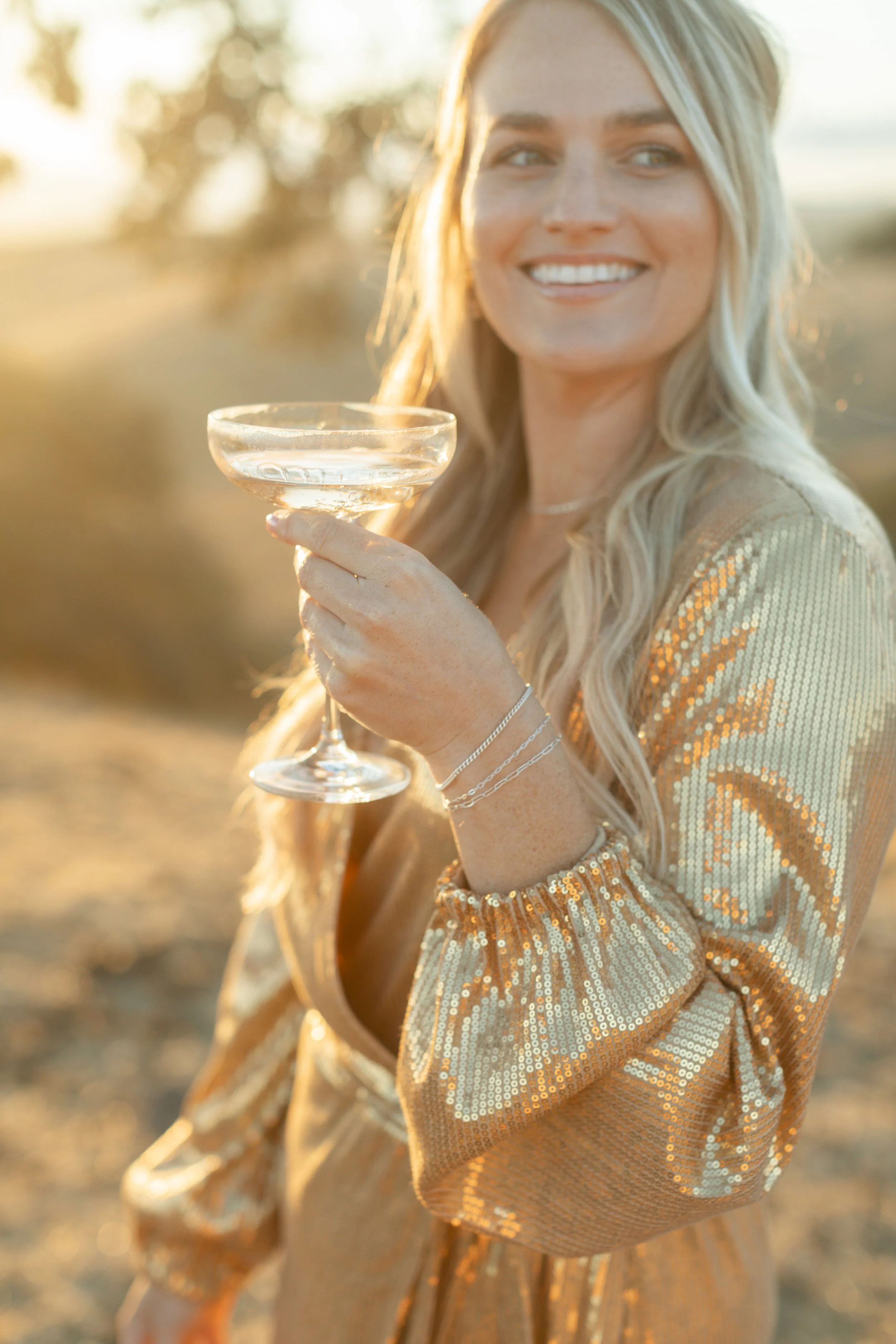 A woman in a sequined gold outfit holding a champagne glass outdoors during sunset, smiling.