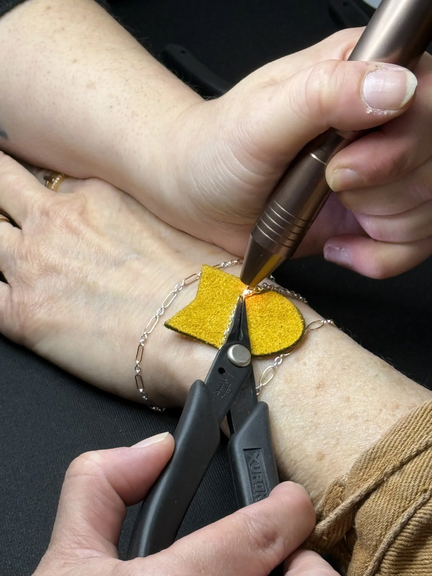 Jewelry piece being engraved on a person's wrist with a handheld engraving tool, while another person holds the jewelry steady with pliers.