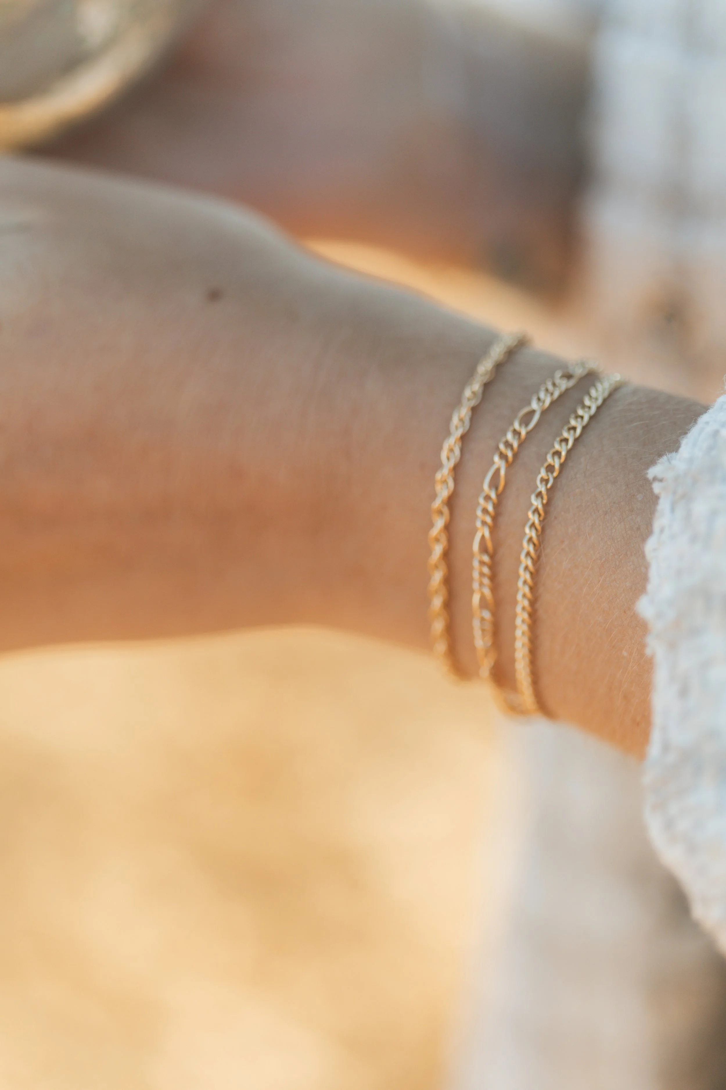 Close-up of a person's arm with three gold bracelets, tan skin, and a white ruffled sleeve.