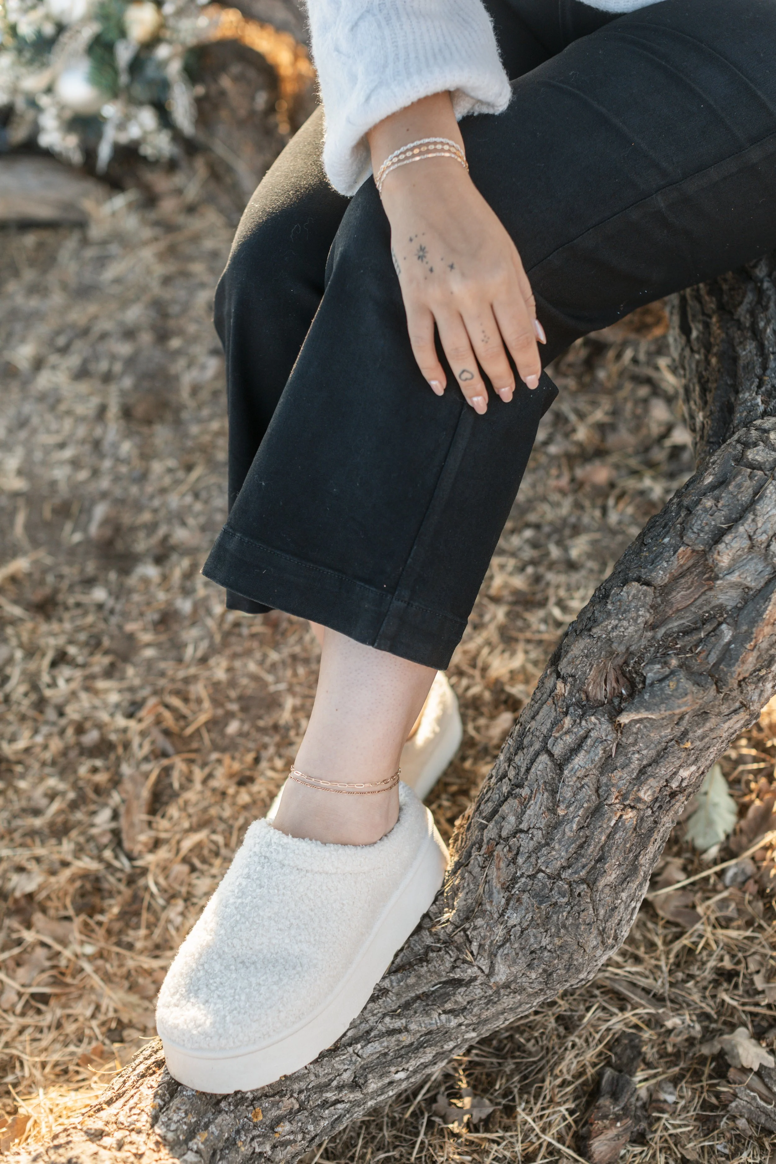 Person sitting on a tree branch wearing white fuzzy slip-on shoes, black pants, a white long-sleeve sweater, and a delicate anklet and wrist bracelets, with autumn leaves on the ground.