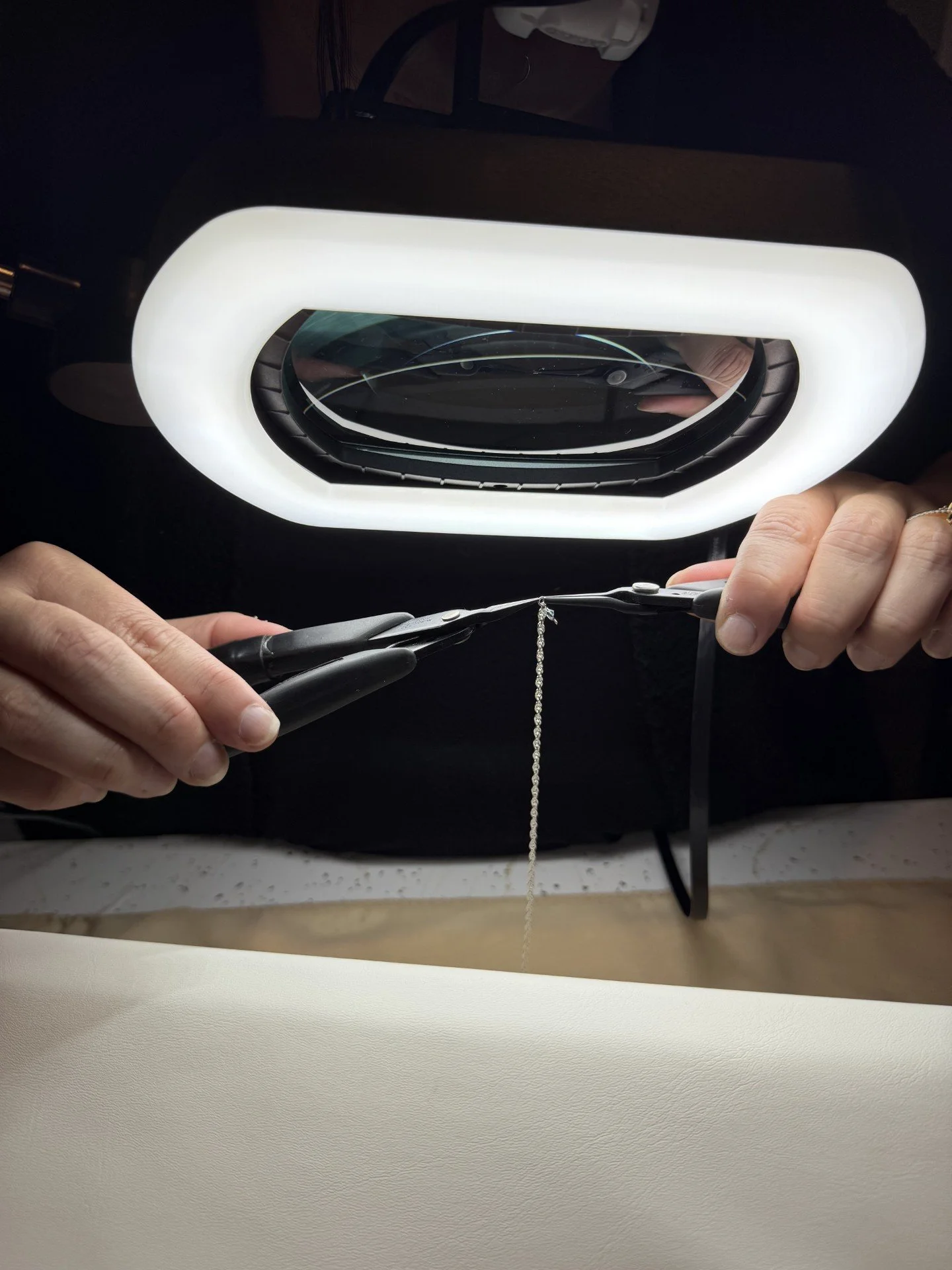 Person's hands working on jewelry, using tools and magnifying lighted lamp for close inspection.