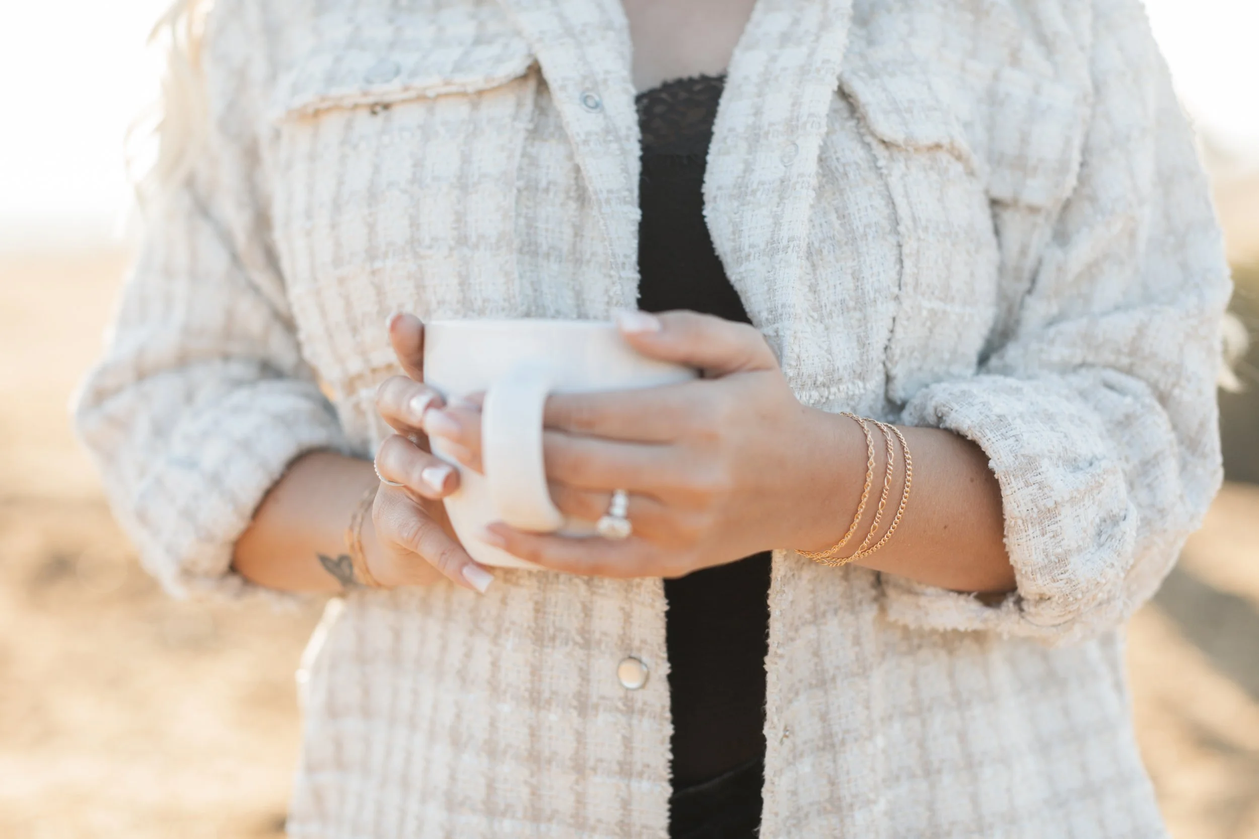 A person wearing a beige textured jacket holds a white mug with both hands, with a tattoo on the wrist and gold jewelry on the wrist, standing outdoors in natural sunlight.