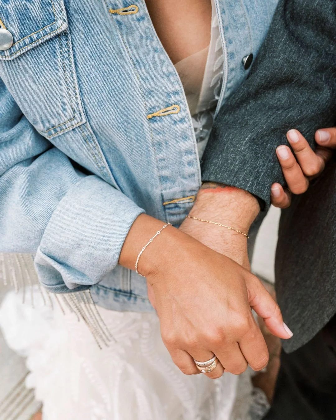 Close-up of two people holding hands, one wearing a light blue denim jacket and the other wearing a dark gray jacket, with focus on their hands and bracelets.
