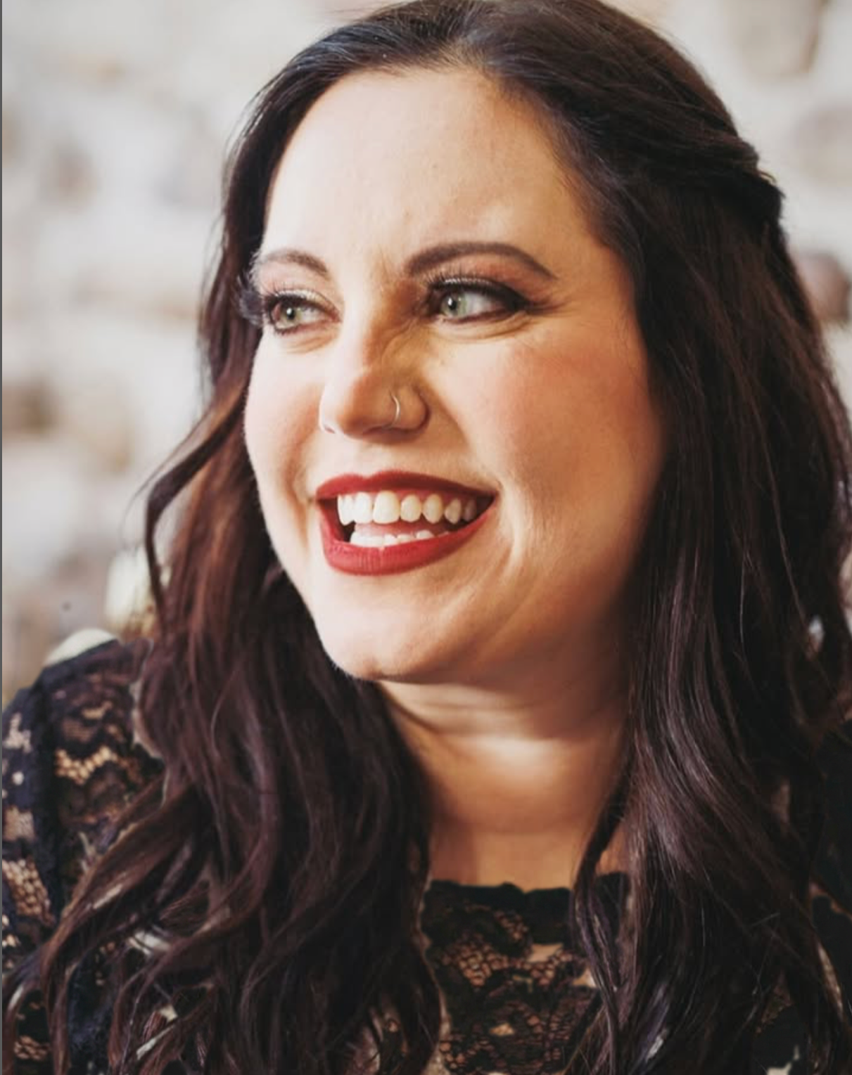 Close-up of Amber, owner of Tesori Collective, with long dark hair, green eyes, and red lipstick, smiling and looking to the side, wearing a black lace top, against a blurred light-colored background.