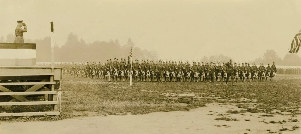 A sepia-toned historical photo of a military parade with soldiers marching in formation on a field, with an American flag and a person standing on a platform observing.