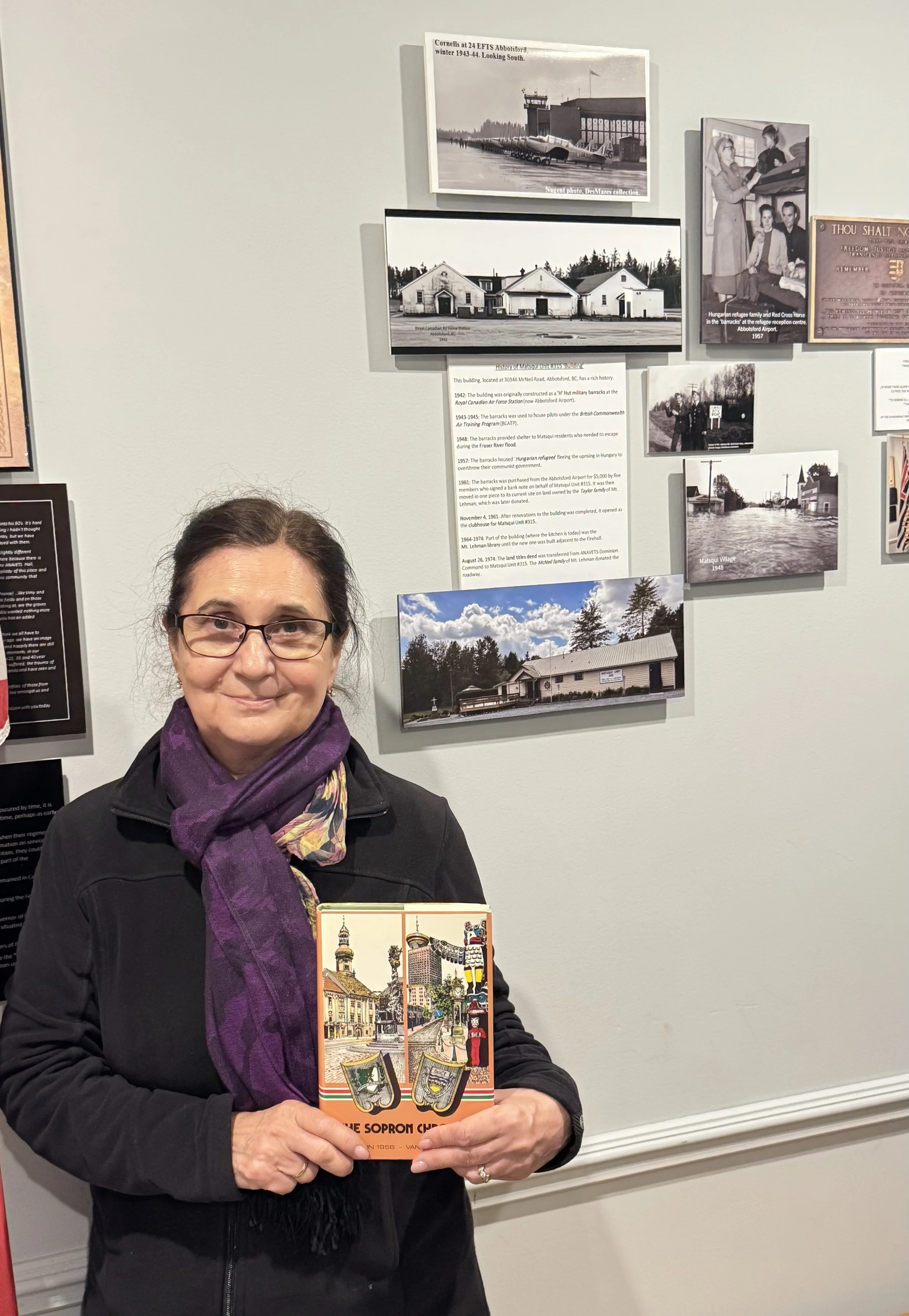 Woman with glasses and purple scarf holding a vintage postcard. She stands in front of a gray wall with historical and black-and-white photographs and informational text about a local history exhibit.