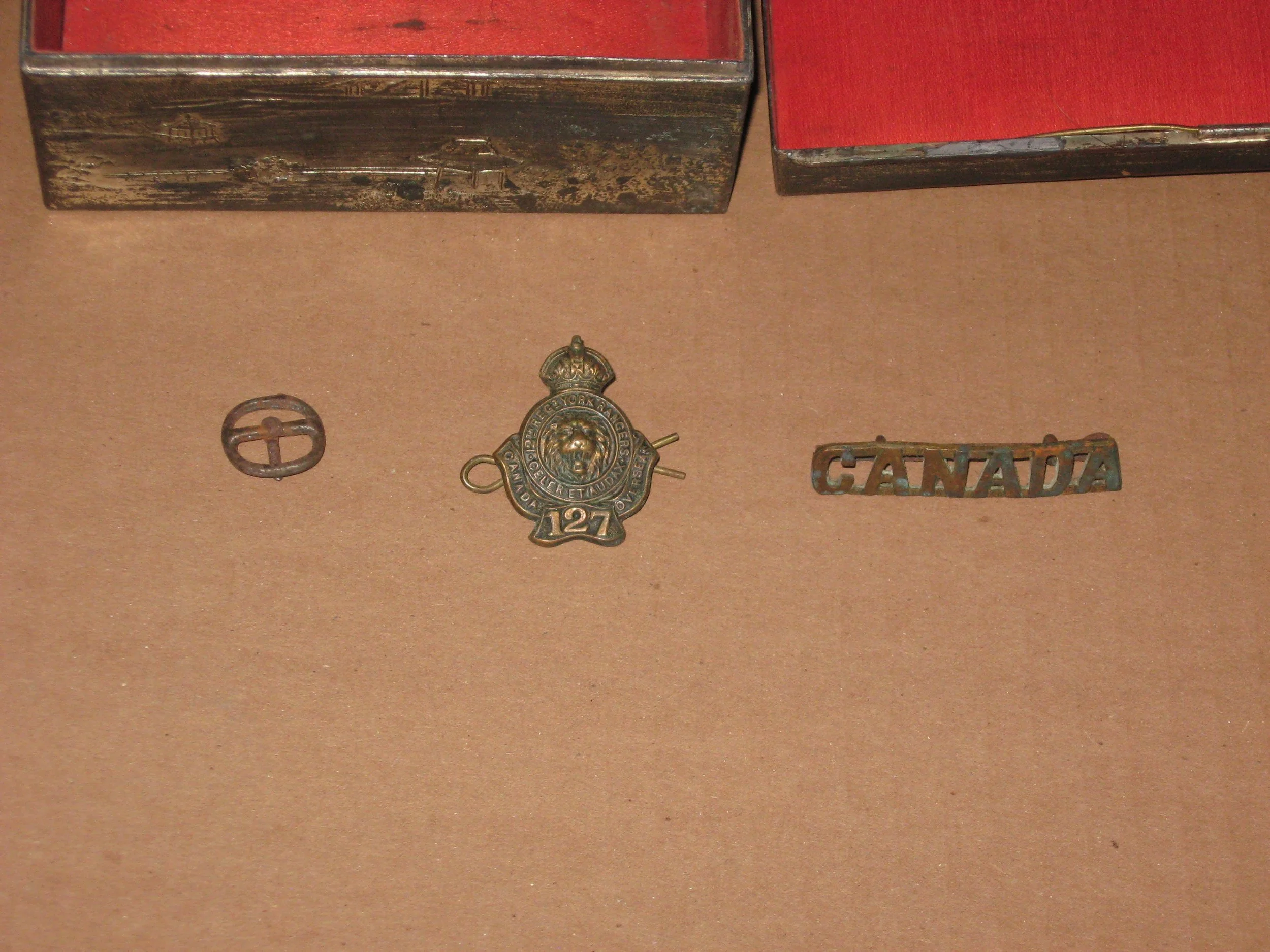 Three small vintage metal objects: a buckle, a badge with a lion and crown, and a cutout spelling 'CANADA', arranged on a brown surface.