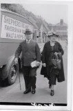 Two people walking on a street, holding cups, with a vintage bus nearby and a sign in the background.
