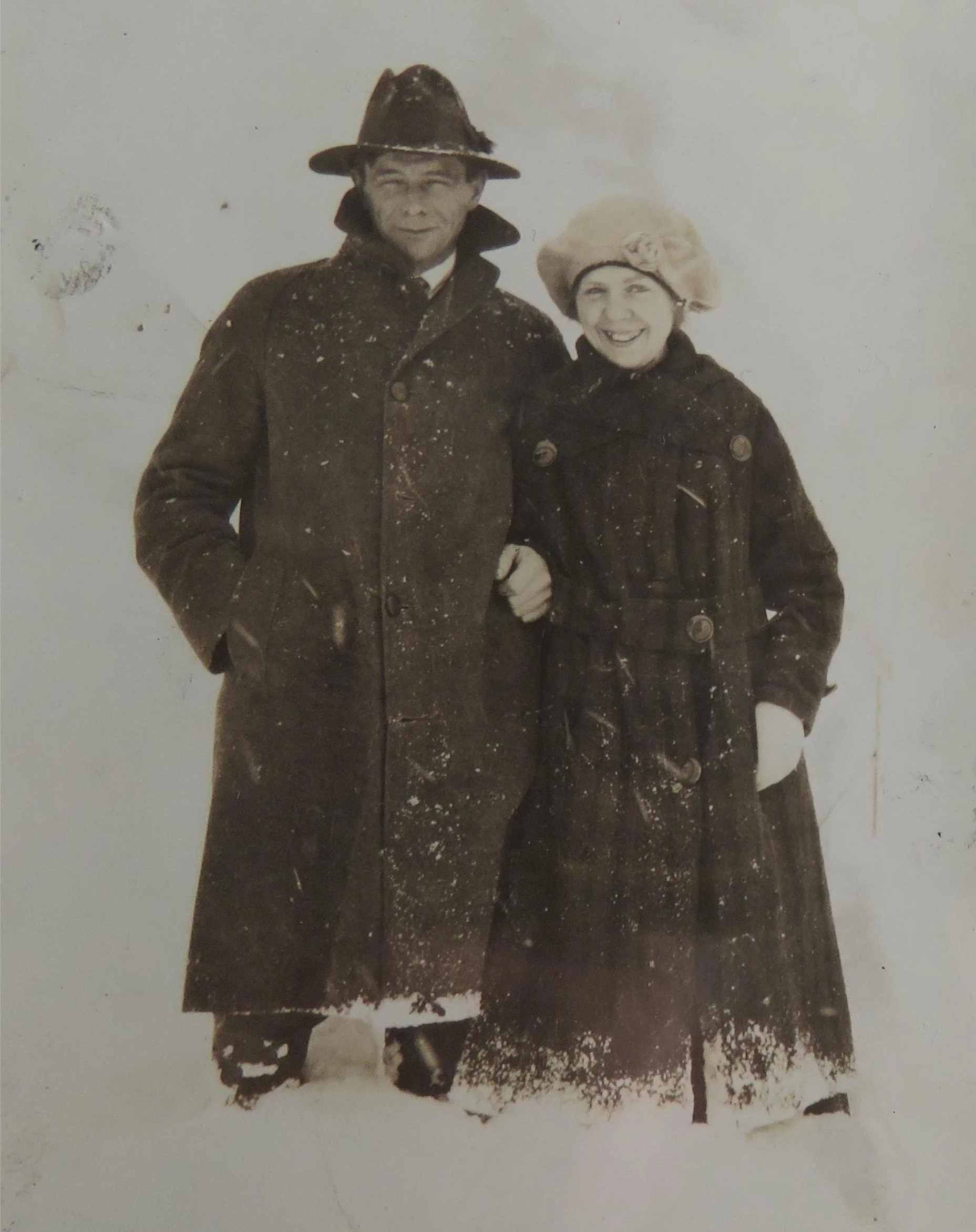An old black-and-white photograph of a man and woman standing in the snow, both wearing heavy winter coats and hats, smiling at the camera.