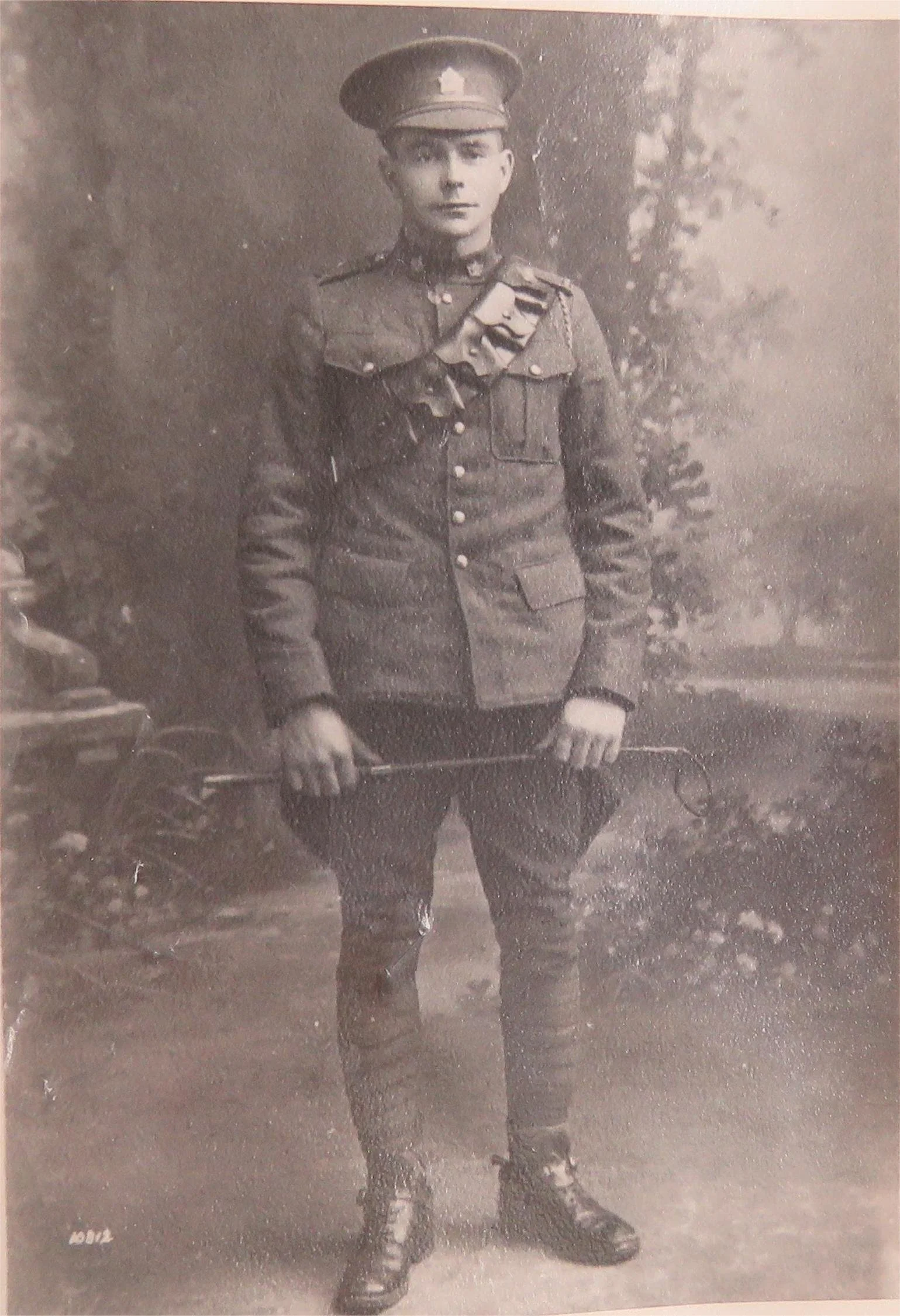 Black and white photograph of a young soldier in a military uniform, standing outdoors with trees in the background, holding a sword or baton in hands.