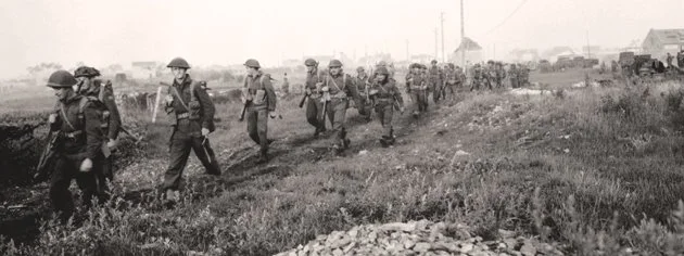 Black and white photo of soldiers marching through a grassy field during wartime, with tents and buildings in the background.