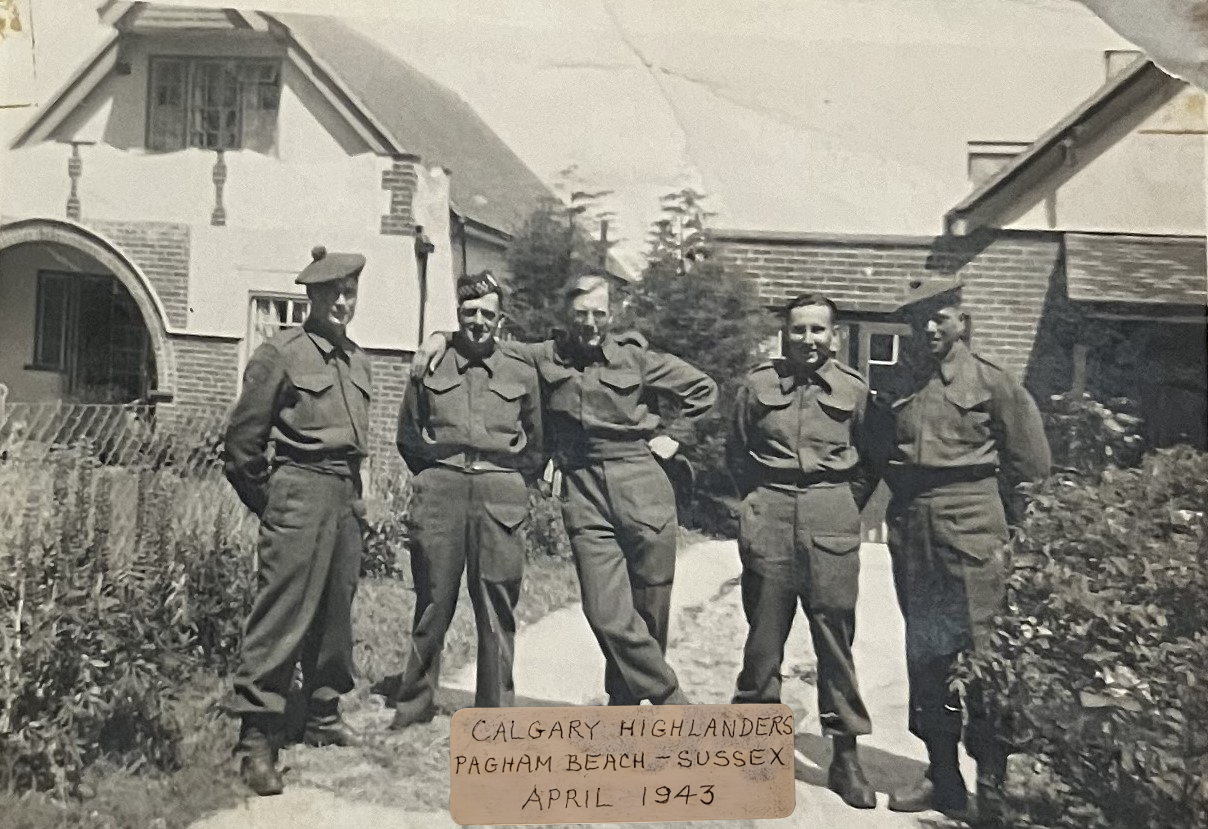 Black and white photograph of five soldiers in uniform standing outdoors on a dirt path, with houses and trees in the background. A handwritten sign in front reads "Calgary Highlanders Pagham Beach - Sussex April 1943."