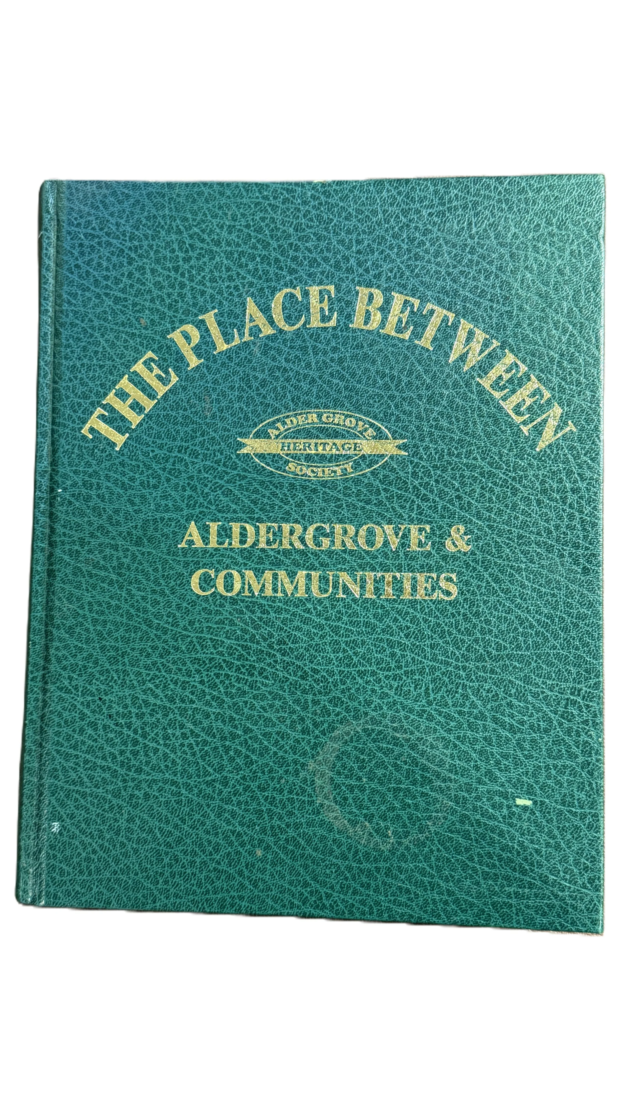 A teal book titled 'The Place Between' by Alder Grove & Communities, with gold lettering and a logo for the Alder Grove Heritage Society.