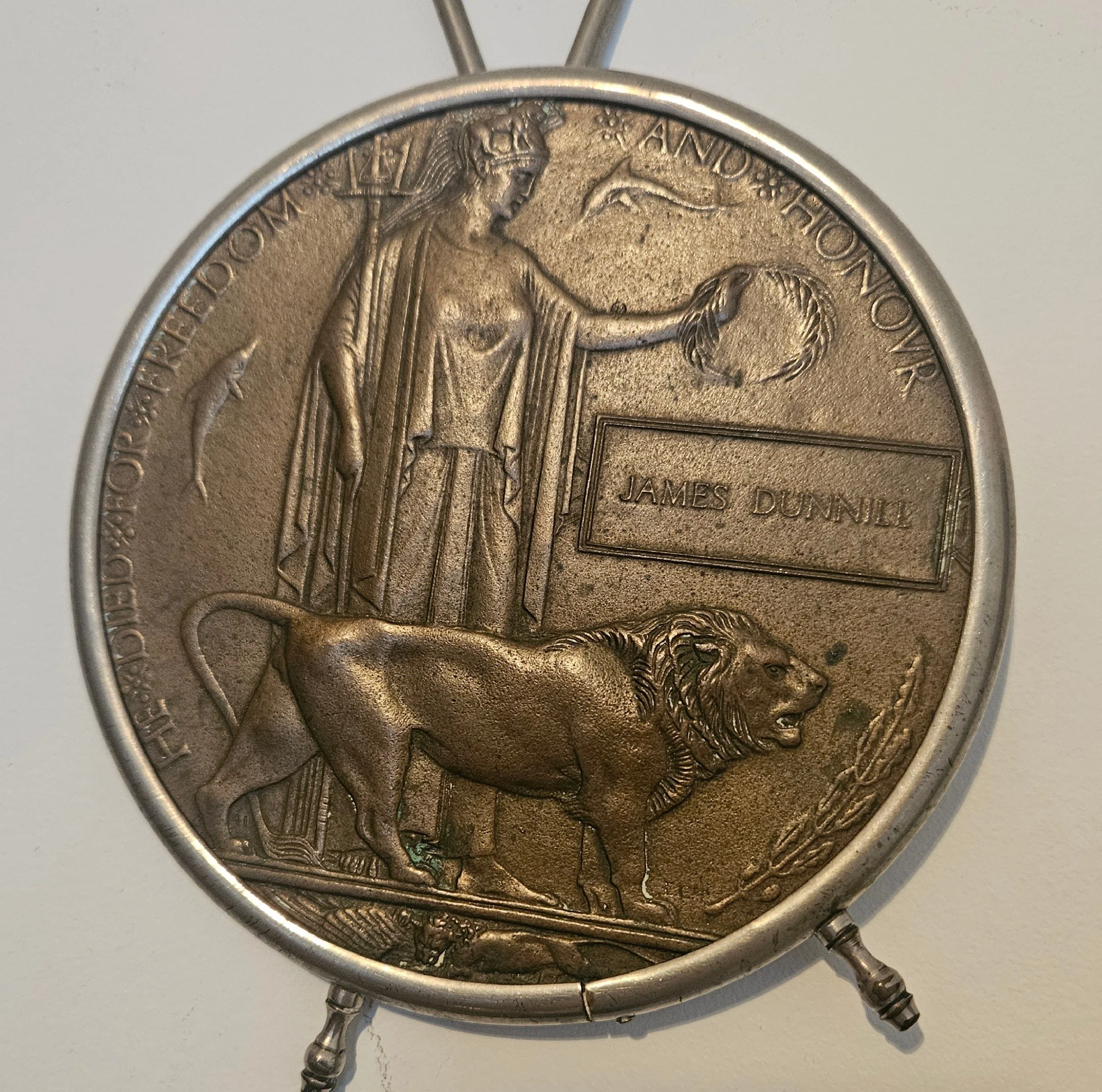 A circular metal coin featuring a design with a woman holding a wreath and a dolphin, a lion standing below, and an engraved plaque with the name James Dunnill.