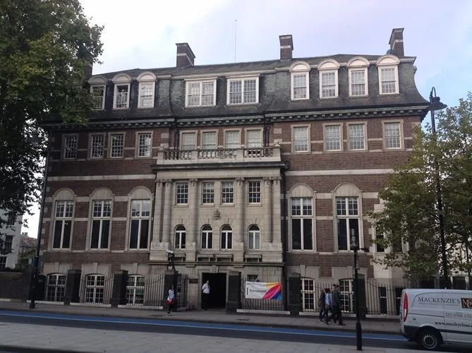 A multi-story historic brick building with large windows, decorative stone accents, and a main entrance with columns and a balcony. People are walking nearby on the sidewalk.