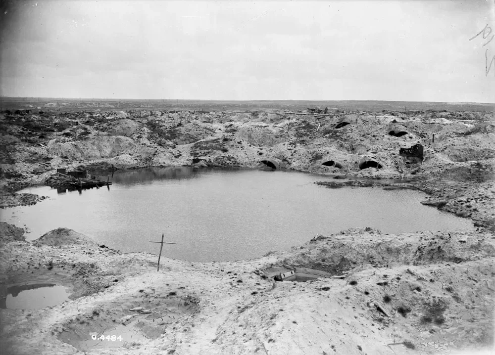 Black and white photograph of a barren landscape with a large water-filled crater in the center surrounded by sandy terrain and multiple ventilation pipes on the far side.