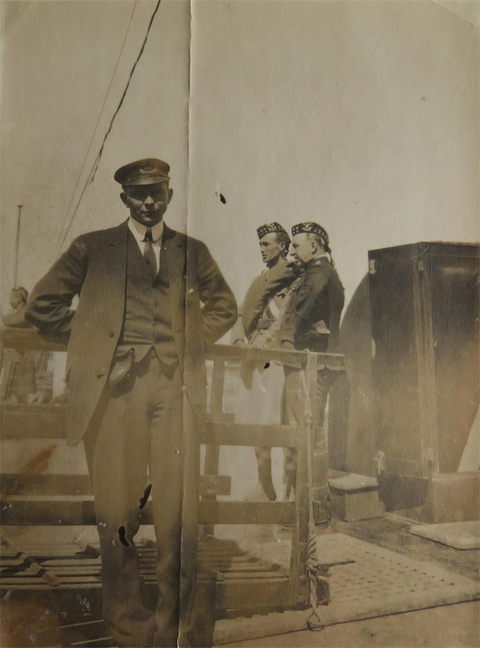 Black and white photograph of four men in military uniforms, one standing in the foreground with hands behind back, and three in the background standing near a railing. The men are wearing hats with military insignia, and the background suggests an o