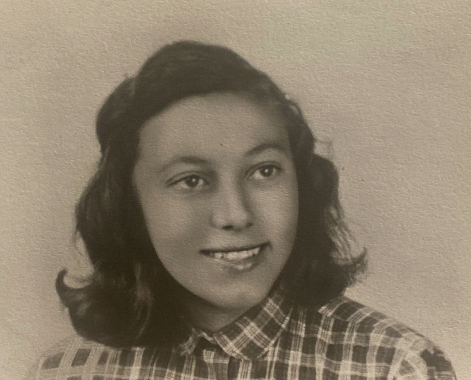 Black and white portrait of a young woman with wavy, shoulder-length hair, wearing a collared, checkered shirt, looking slightly to the side and smiling.