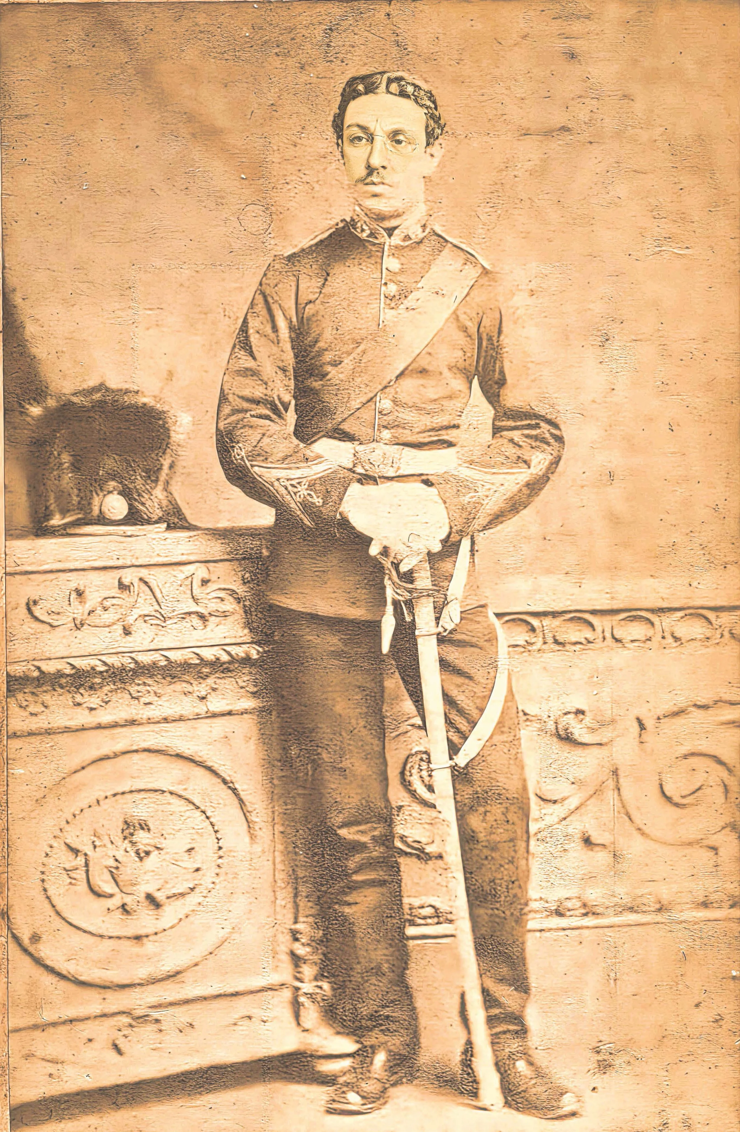 Sepia-toned vintage photograph of a young man in a military uniform holding a sword, standing next to an ornate wooden piece of furniture with decorative carvings and a fur hat on top.