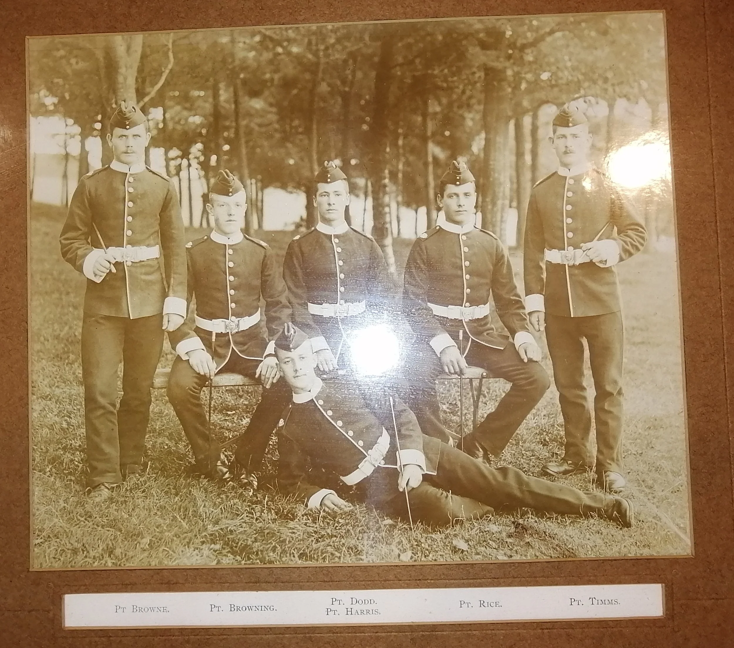 A vintage sepia photograph of six soldiers in uniform outdoors, with five standing and one sitting on the ground, in a grassy area with trees in the background. The soldiers wear military jackets, hats, and belts. There is a nameplate below the photo
