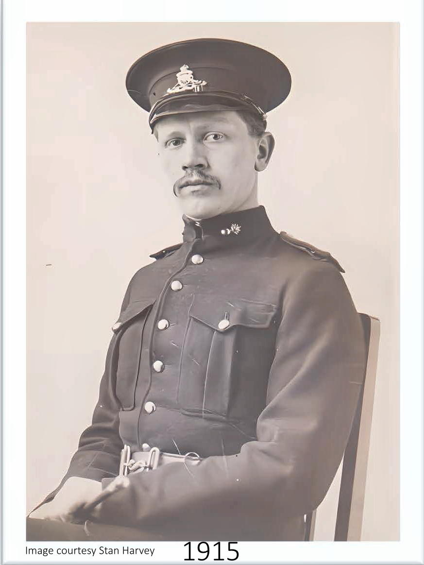 A black and white photograph of a man in a military uniform from 1915, wearing a cap with a badge, sitting on a chair, with a serious expression.