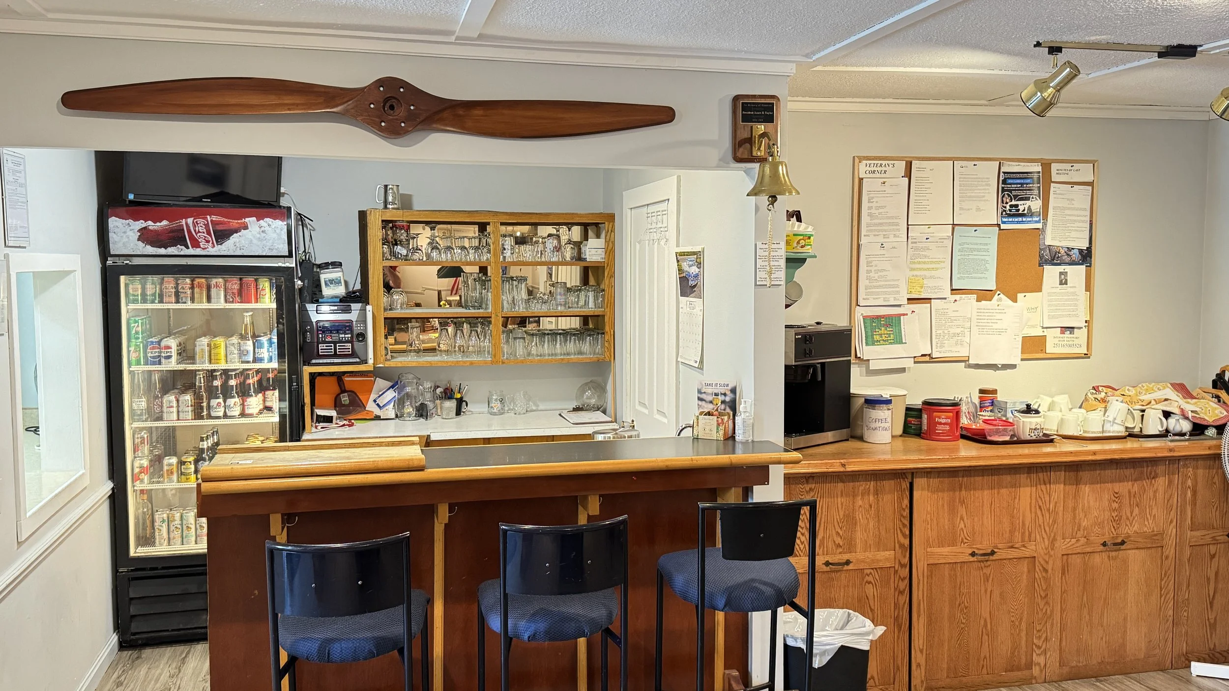 Interior of a small cafe or break room with a wooden counter, bar stools, a refrigerator stocked with drinks, a glassware shelf, and contactless payment terminal. Cork bulletin board with notices and a coffee station.
