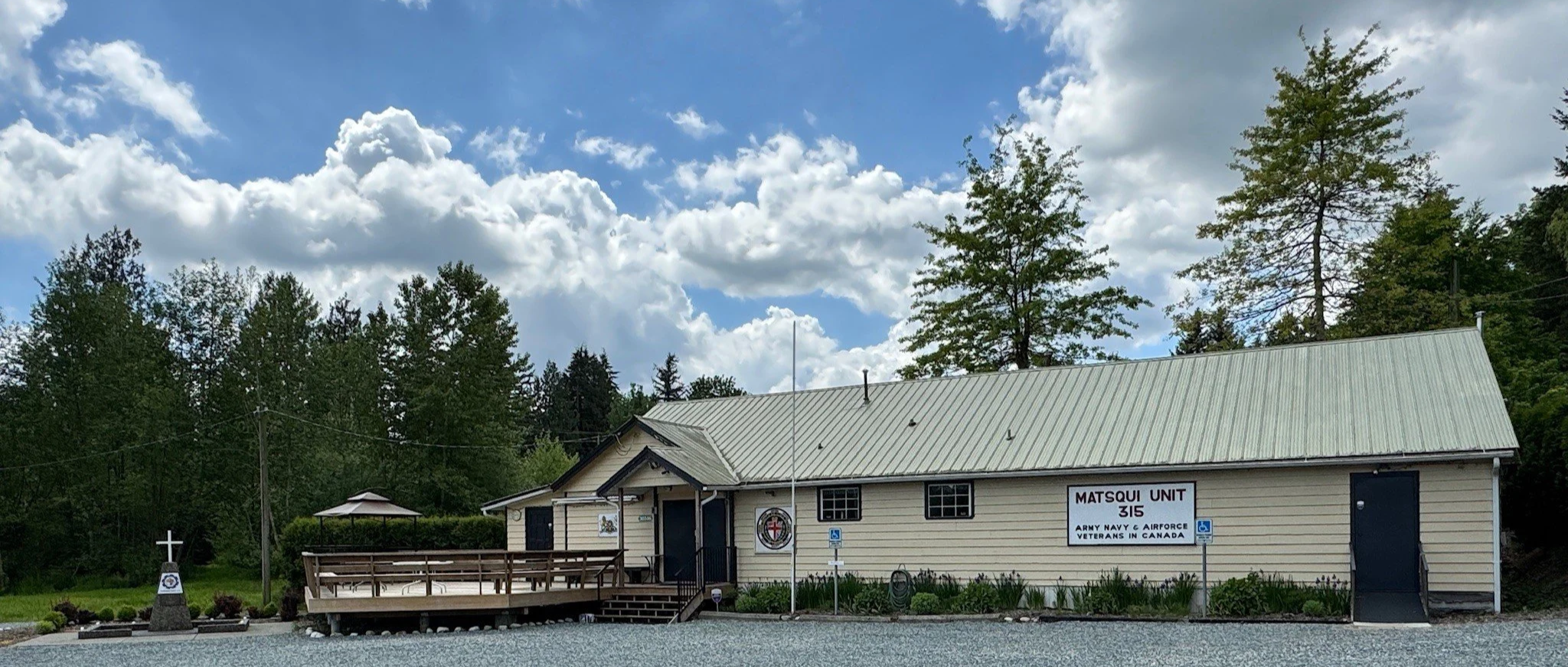 A beige building with a green metal roof labeled 'Matsqui Unit, 315, Army Navy & Airforce Veterans in Canada.' The building has a small porch with stairs, a wheelchair accessible parking sign, and is surrounded by trees and a gravel parking area. There is a monument with a cross and a circle emblem on a stone base in front of the building, with landscaping around it. The sky above is partly cloudy.
