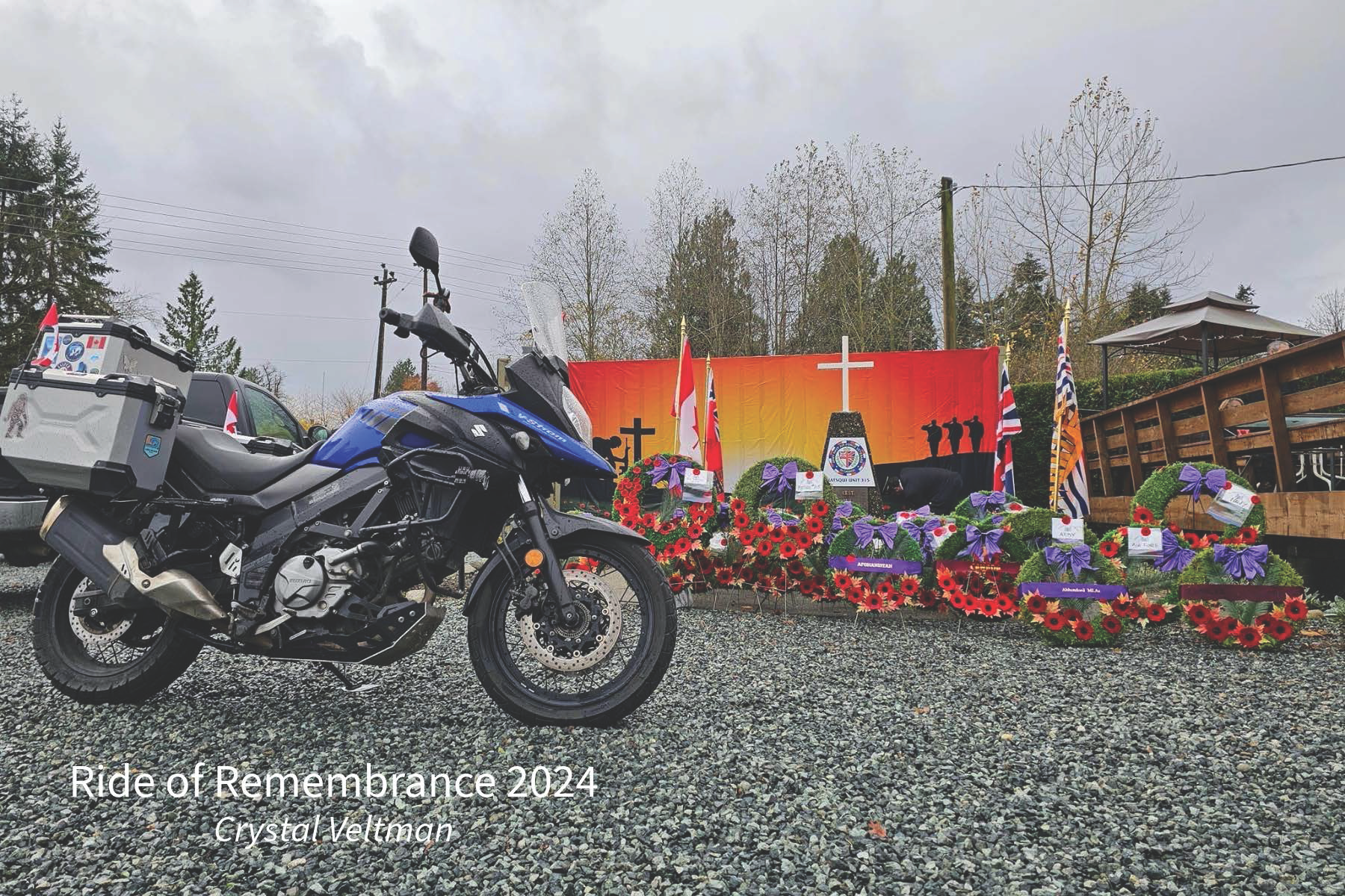A motorcycle parked on a gravel surface in front of a memorial display with red and purple floral arrangements, flags, and a large banner with a Christian cross. The memorial commemorates the Ride of Remembrance 2024, organized by Crystal Veltman.