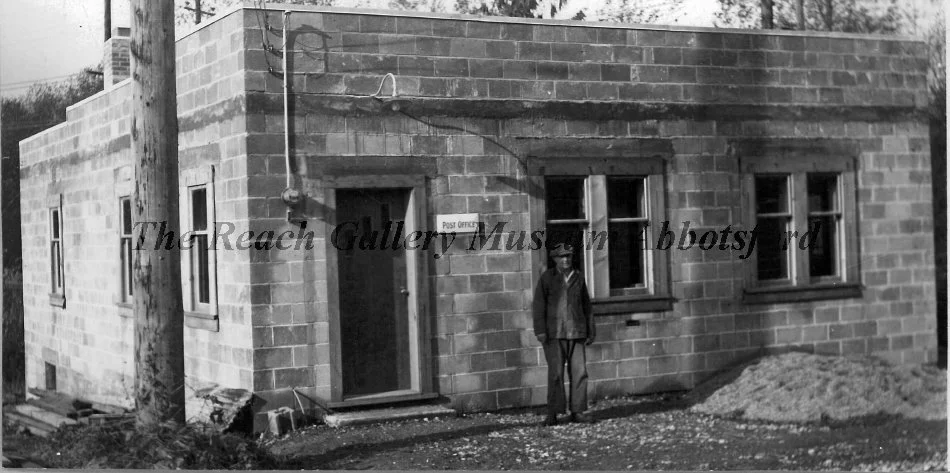 A black and white photo of a brick building with a small person standing outside. The building has three windows and a door with a sign that says 'Post Office.' There is a wooden utility pole in front of the building and a pile of dirt nearby.