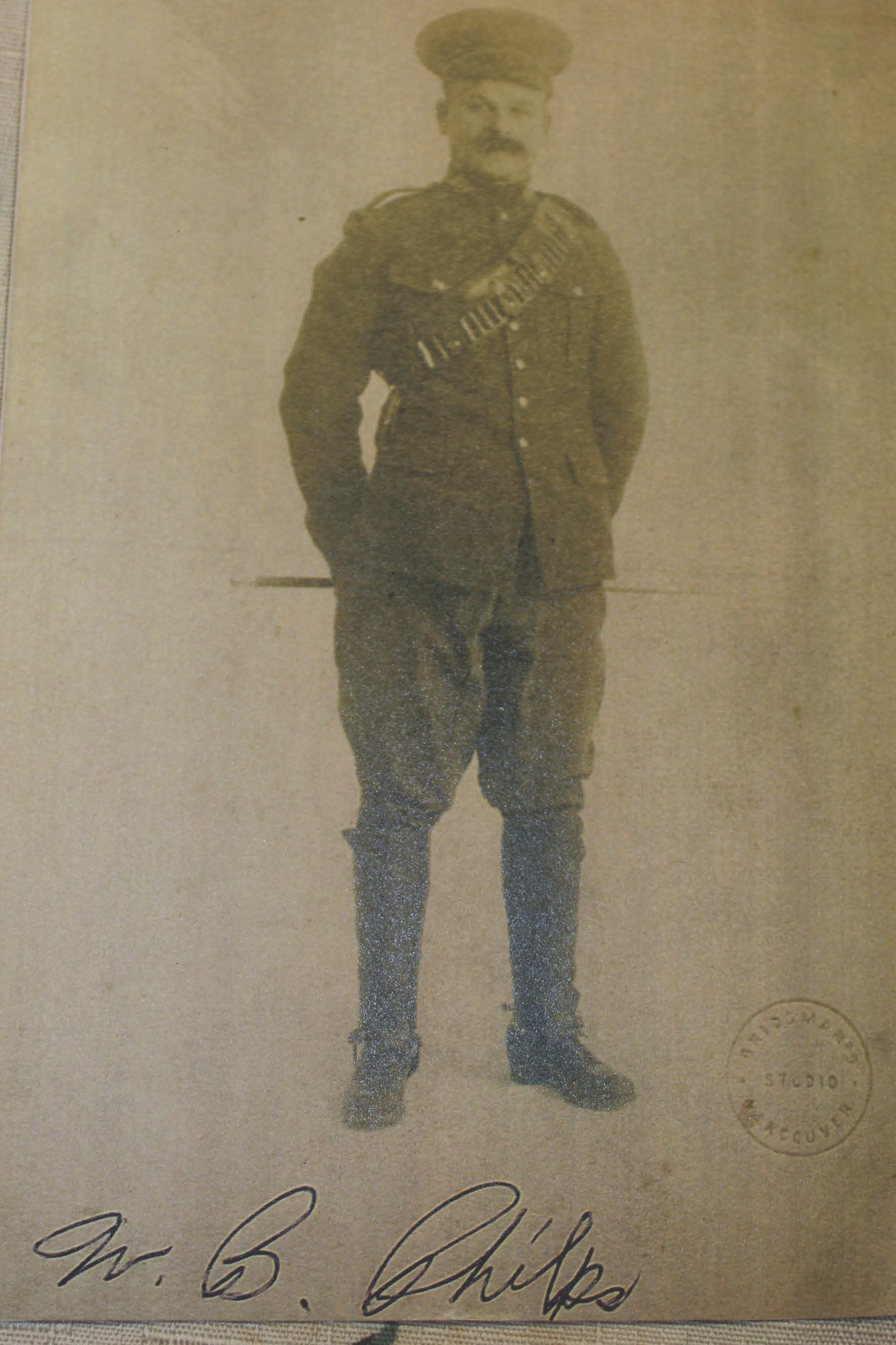 A sepia-toned photograph of a man in a military uniform standing with his hands behind his back, wearing a cap and a bandolier across his chest, with handwritten text and a circular seal in the lower right corner.