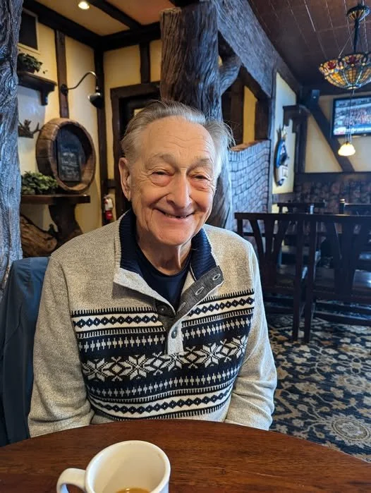 Smiling elderly man in a cozy restaurant with rustic wood decor, sitting at a table with a coffee mug in front of him.