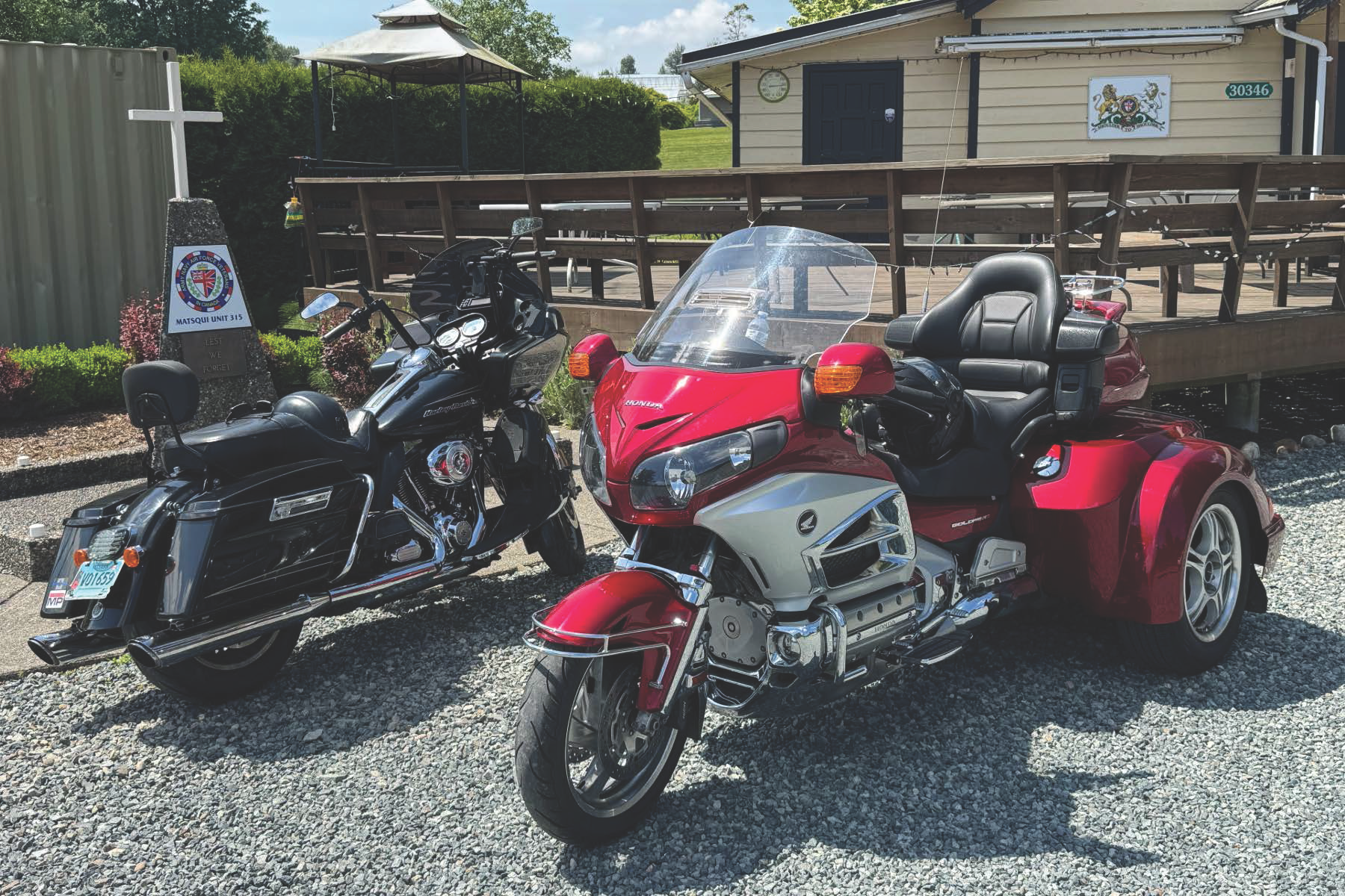 A red and white trike motorcycle parked next to a black touring motorcycle on a gravel surface, with a building, greenery, and a sign in the background.