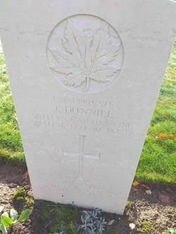 A white gravestone with a carved maple leaf at the top, engraved with the name Johnnie, and a cross below. The gravestone is outdoors on grass.