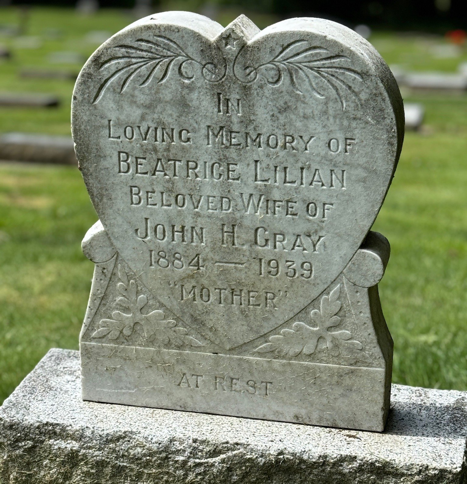 Heart-shaped tombstone with engraved flowers and inscription honoring Beatrice Lillian Gray, wife of John H. Gray, who lived from 1884 to 1939, located in a cemetery with other tombstones in the background.