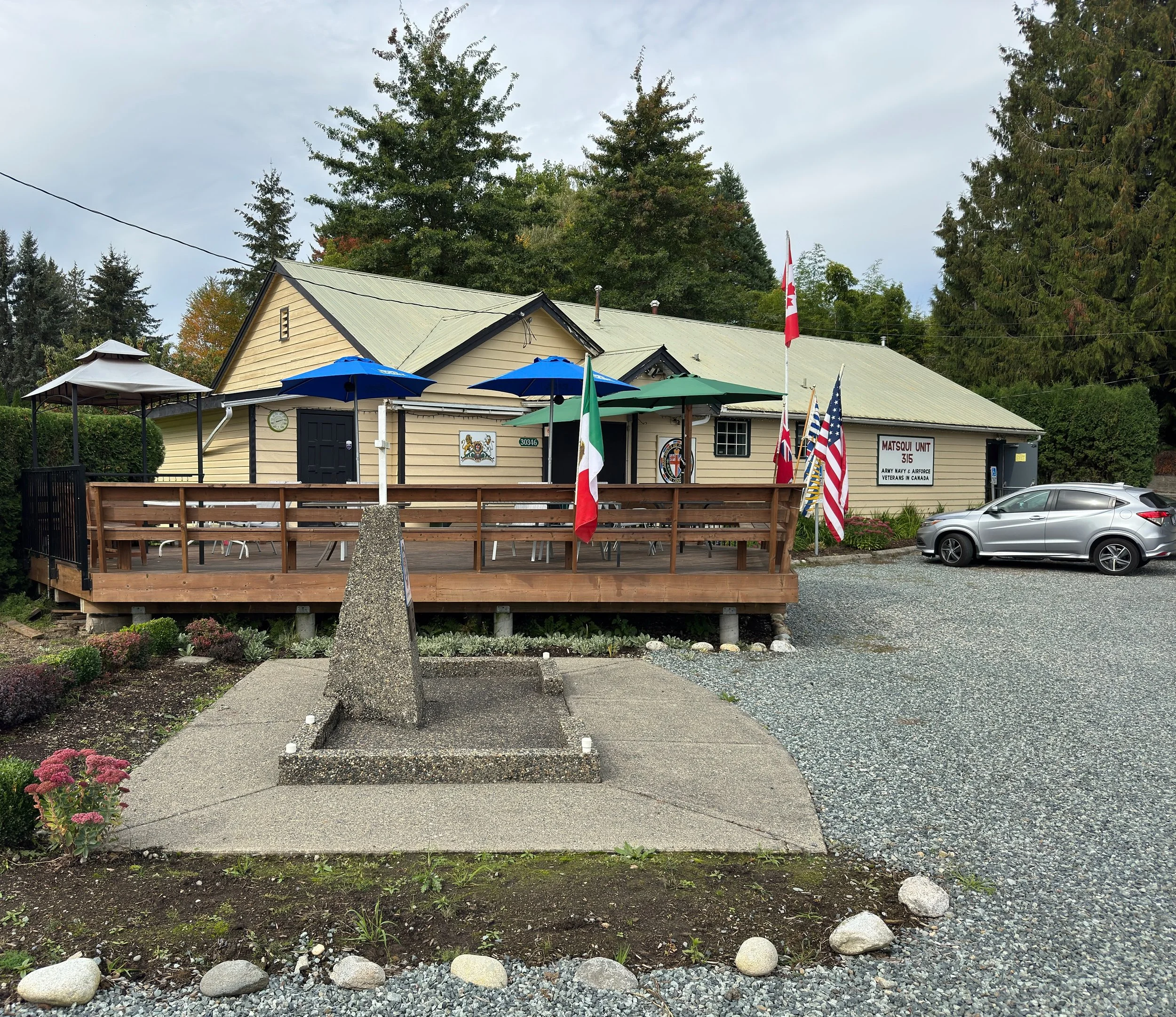 A small building with a porch decorated with flags, umbrellas, and signs. There is a parking lot with a silver car, and surrounding greenery with trees.