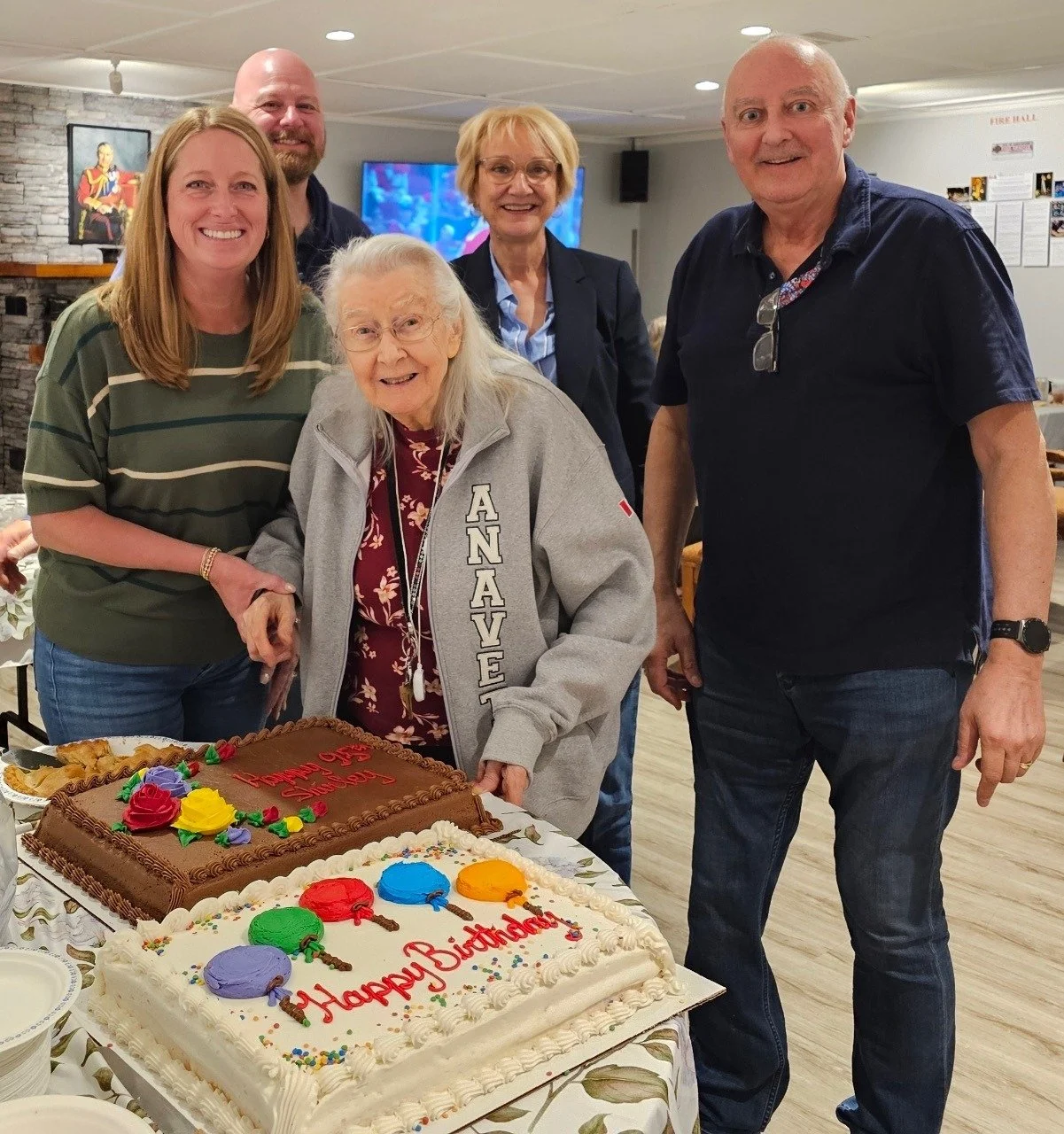 Group of six people celebrating a birthday with two cakes, including an elderly woman in a gray jacket holding a cake, all smiling in a dining area.