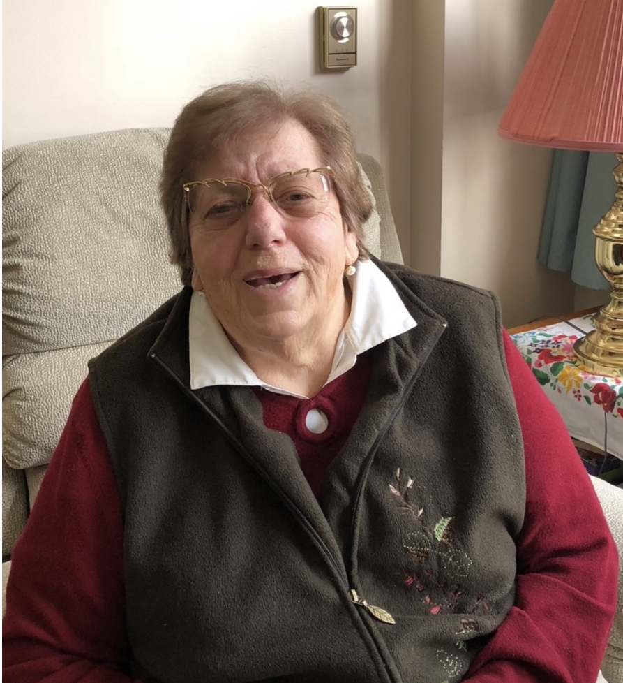 A smiling elderly woman with short gray hair, glasses, and pearl earrings, sitting in a beige armchair. She is wearing a maroon sweater, a white collared shirt, and a dark vest. There is a pink lampshade and a table with a colorful cloth behind her.