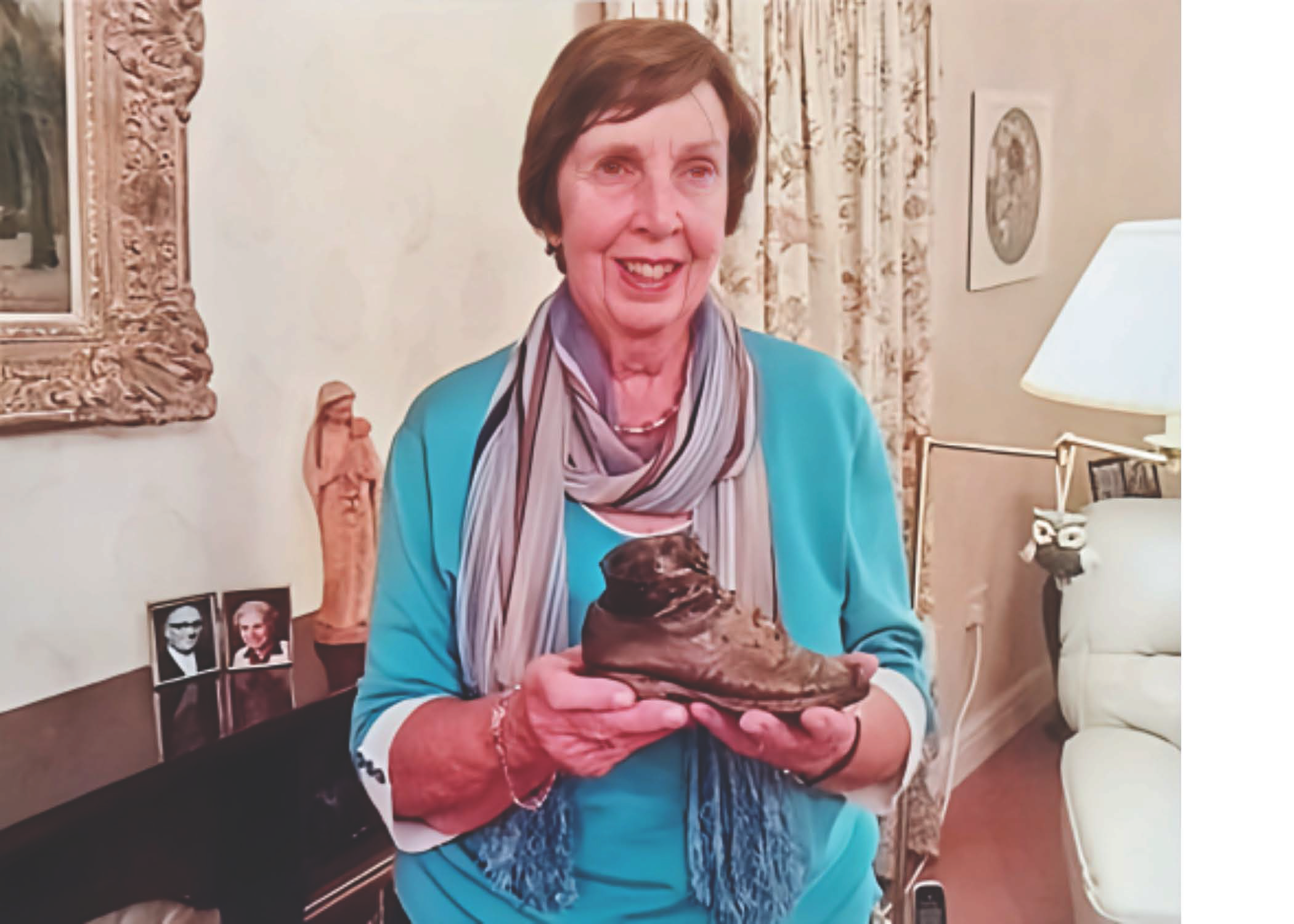 An elderly woman with short brown hair smiling and holding a large ice cream sundae in a glass bowl. She is sitting in a cozy, well-lit room with decorated curtains and framed photographs on a leading surface behind her.