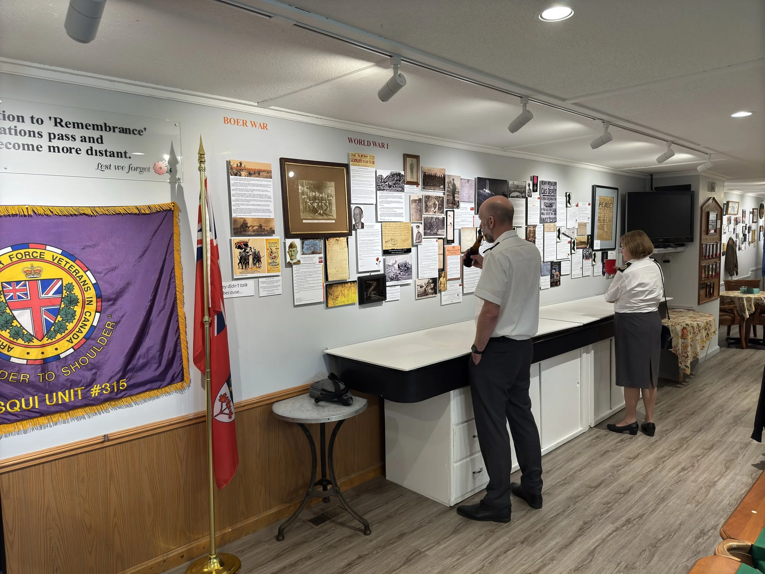 Museum exhibit featuring WWII and Vietnam War displays, with a large flag on the left and two visitors, a man and a woman, viewing informational panels on the wall.