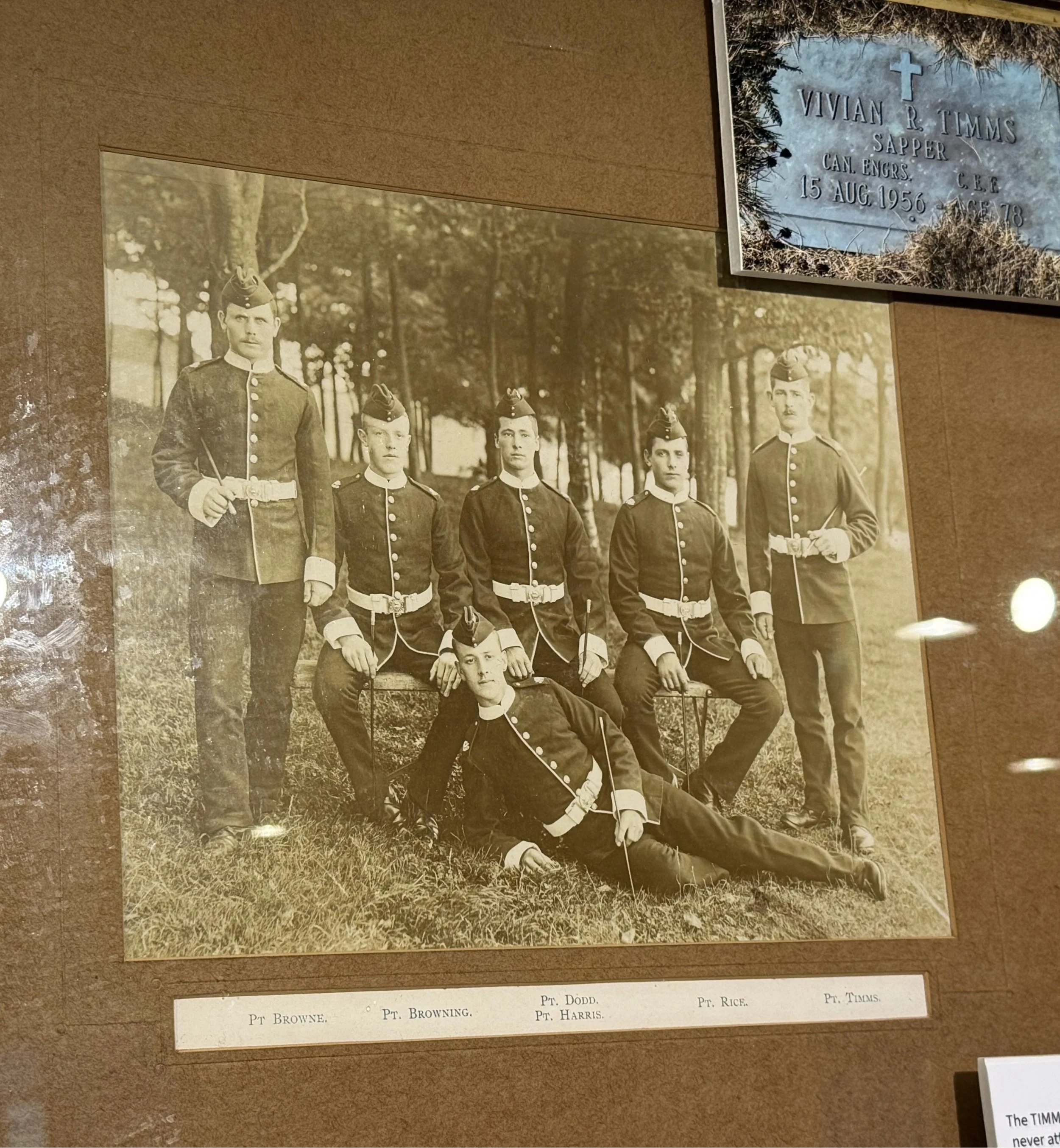 A sepia-toned vintage photograph of seven soldiers in uniform posing outdoors, with trees in the background. One soldier is lying on the ground in front, and the others are standing or seated around him.