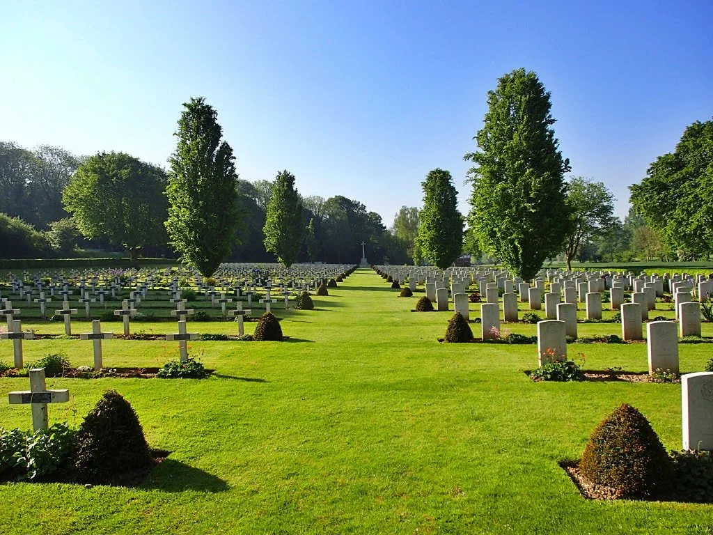 A cemetery with evenly spaced white crosses and upright headstones, well-manicured grass, and tall trees in the background under a clear blue sky.