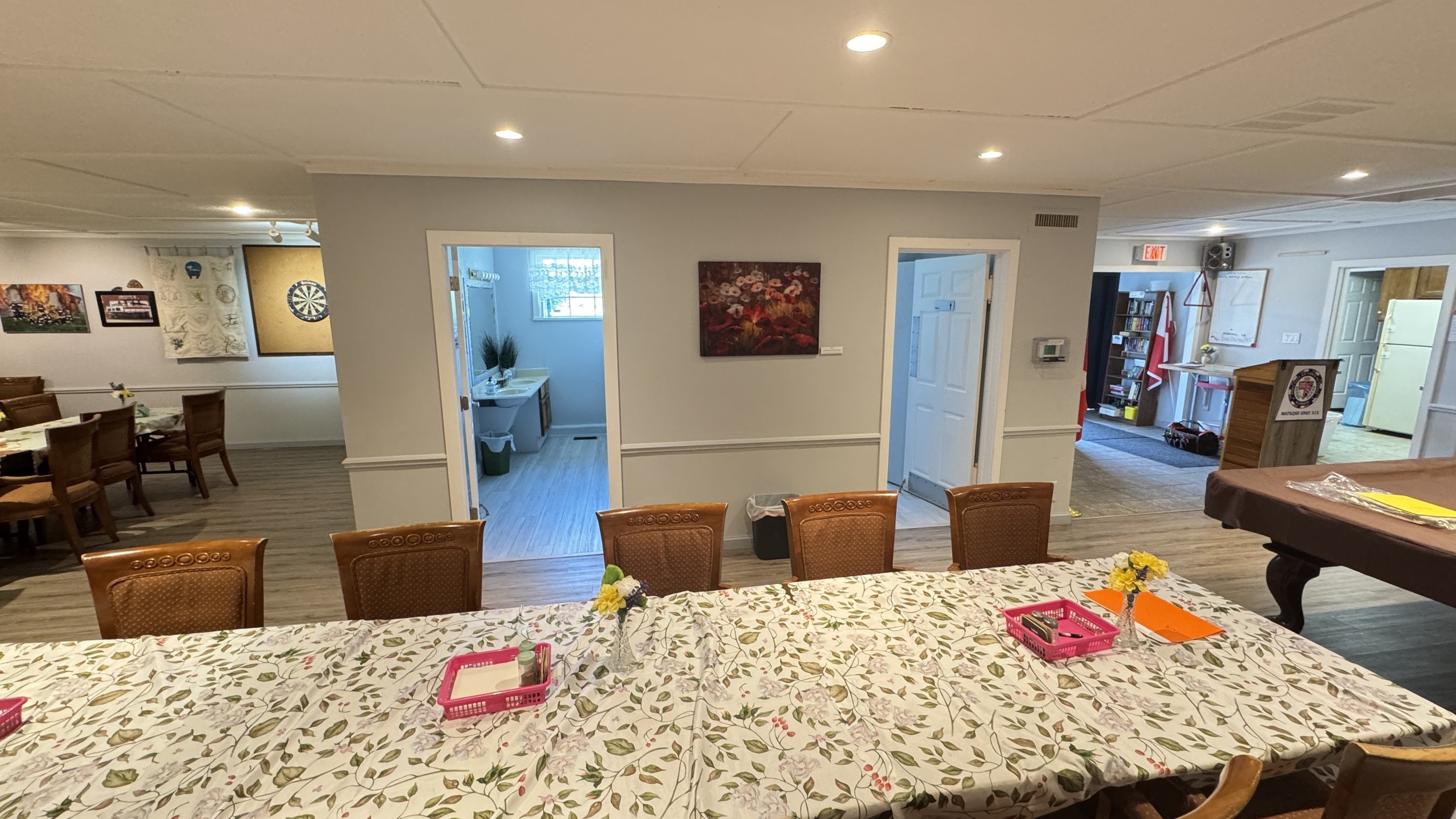 A large dining table covered with a floral tablecloth, surrounded by wooden chairs, with small flower vases and pink and orange papers on it. The room has a wood floor and walls decorated with artwork, including a dartboard. There are multiple rooms and hallways visible, with a sink area in the background and a separate section with a whiteboard, bookshelf, and a pool table.