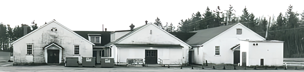 A row of old white buildings with pitched roofs, some with visible doors and windows, situated in a rural or semi-rural area with trees in the background.