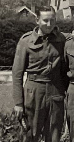 Black and white photo of a man in military uniform standing outdoors, smiling, with a house and shrubbery in the background.