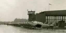 Black and white photo of a row of military airplanes parked on an airfield near a control tower.