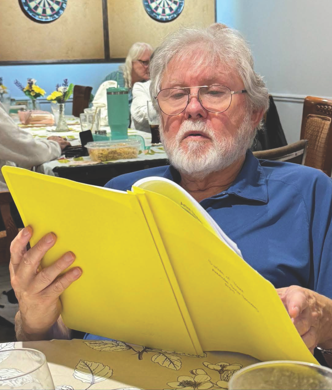 An elderly man with glasses and a beard, wearing a blue shirt, reads a large yellow notebook or book, sitting at a table in a restaurant or dining area, with other people in the background, some with long white hair.