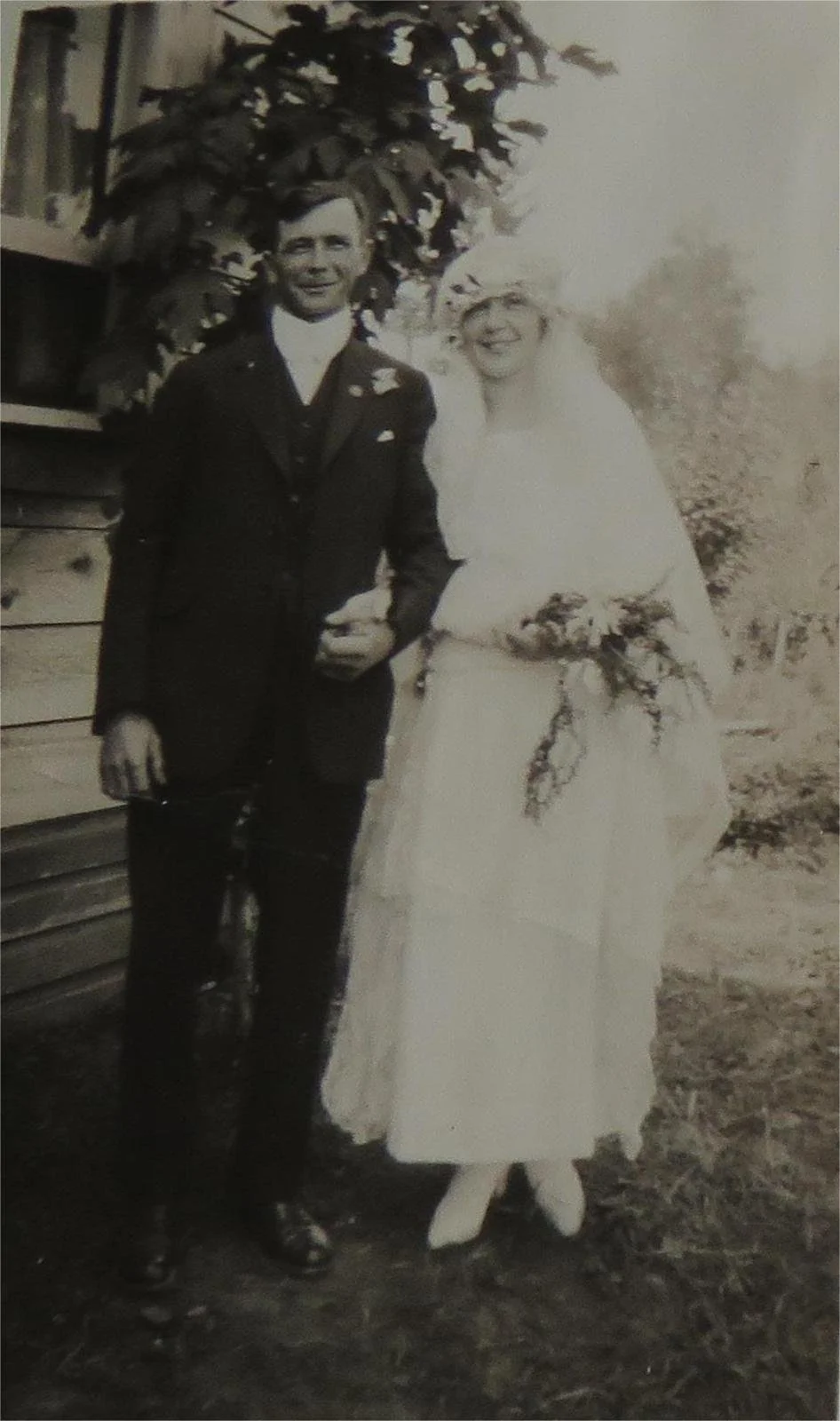 A black-and-white photo of a couple on their wedding day, with the groom in a dark suit and the bride in a white wedding dress holding a bouquet, standing outdoors near a house and a tree.