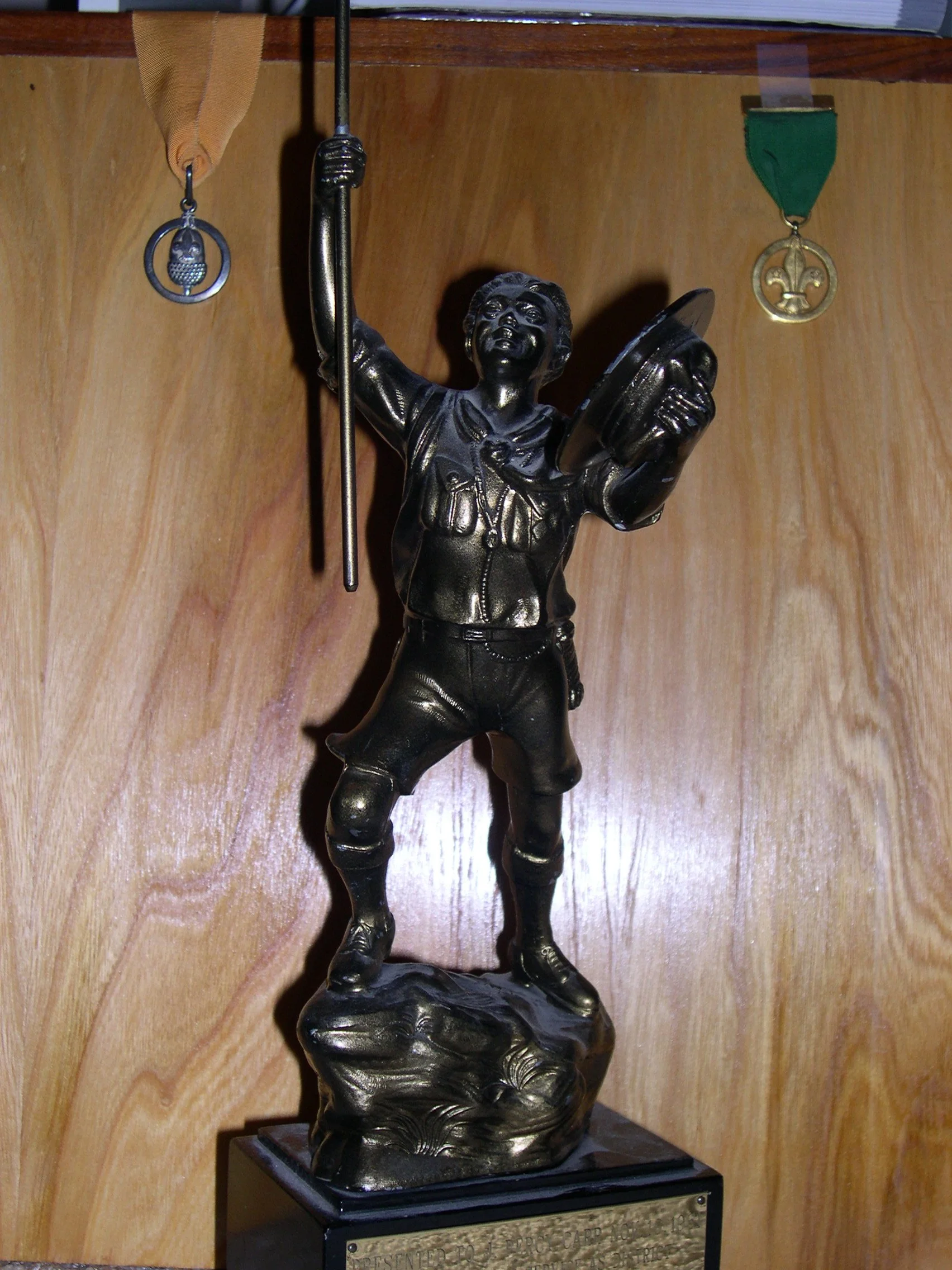 A bronze-colored figurine of a boy scout holding a flagpole and a book, standing on a rocky base, with two medals hanging on the wall behind it.