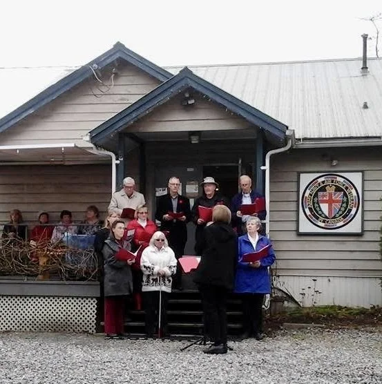 A group of people singing outdoors in front of a house with a flag and coat of arms.