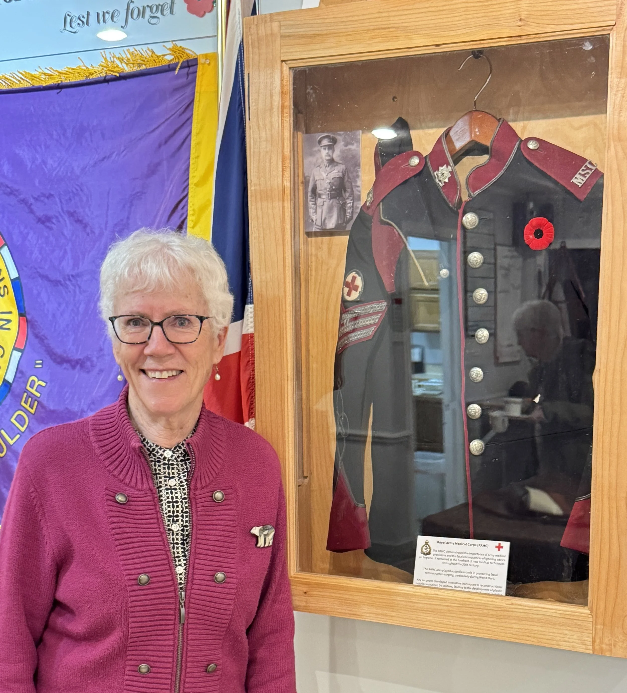 A smiling elderly woman with glasses and a pink cardigan standing next to a display case containing a vintage military uniform with various insignia and a red poppy pin, with a purple and yellow banner and American flag in the background.